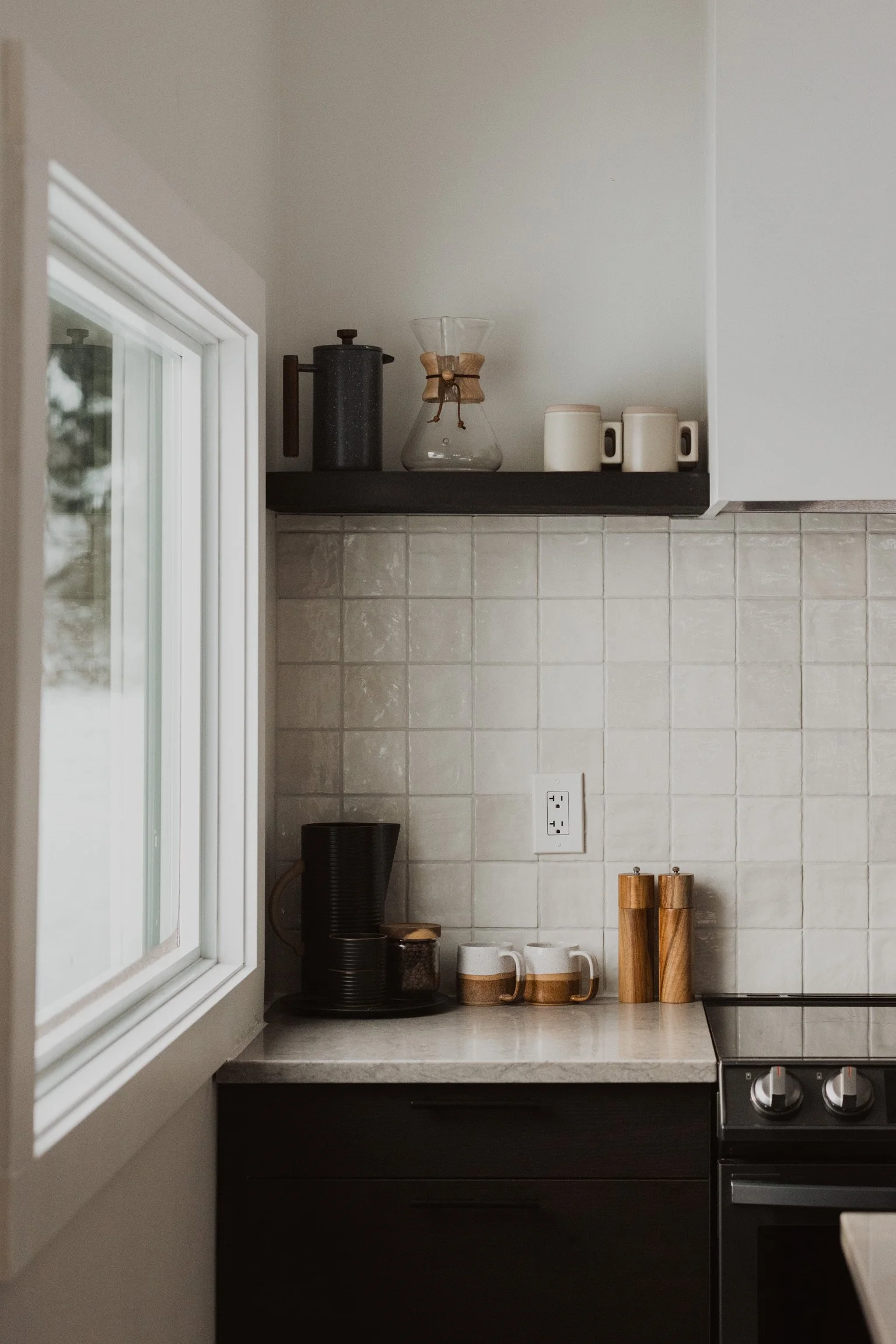 A section of a kitchen counter with coffee mugs, pepper and salt shakers, a coffee grinder, and a window on the left side. Above the counter, a shelf holds coffee carafe, glass coffee brewing device, and containers. The wall is covered with white tiles.