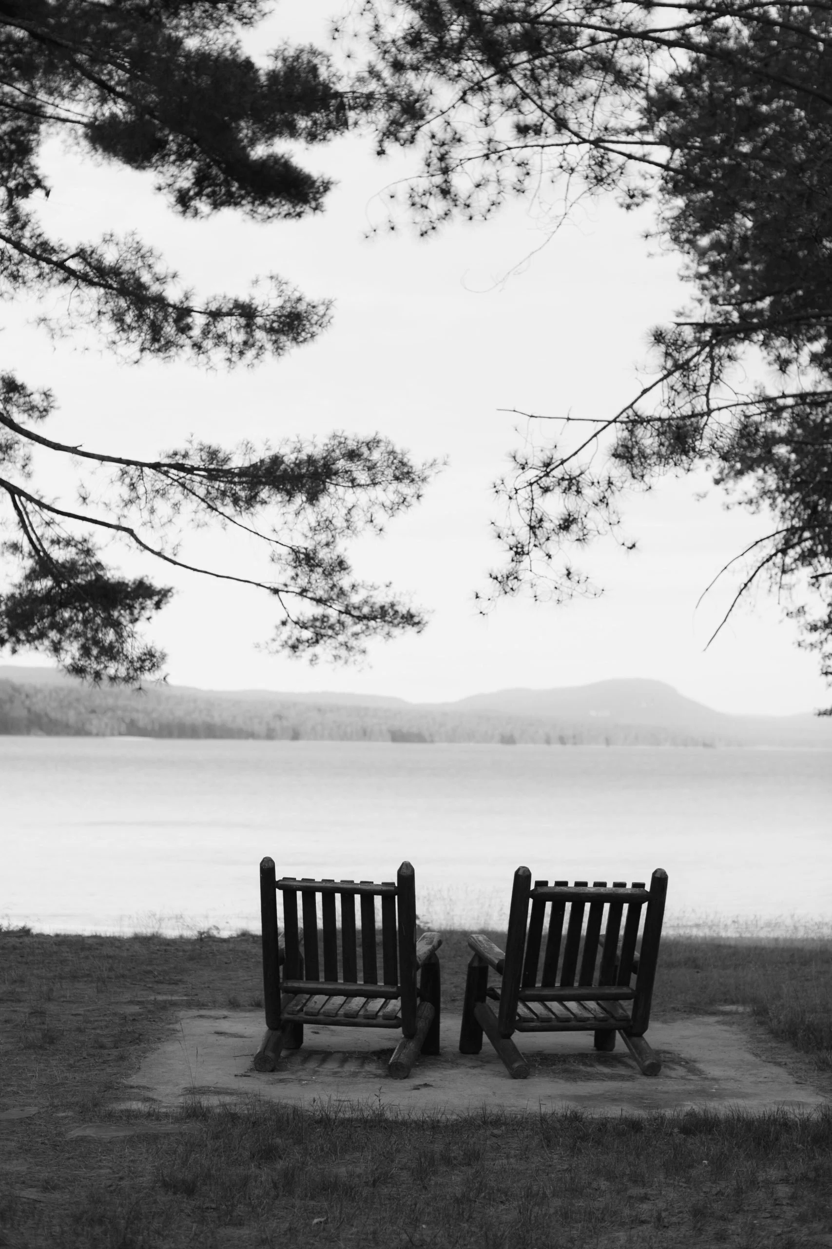 Two wooden chairs facing a lake with mountains in the distance, framed by tree branches overhead.