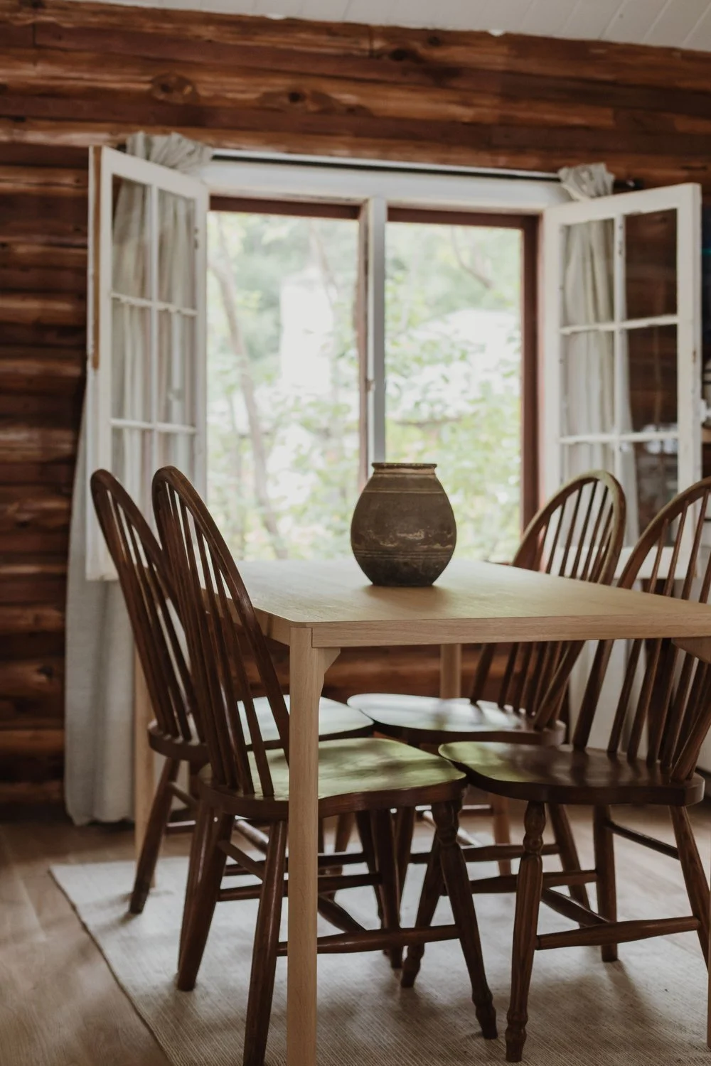 Interior of a rustic dining room with a wooden table, six wooden chairs, a brown vase on the table, an open window with white curtains, wood-paneled walls, and a view of trees through the window.