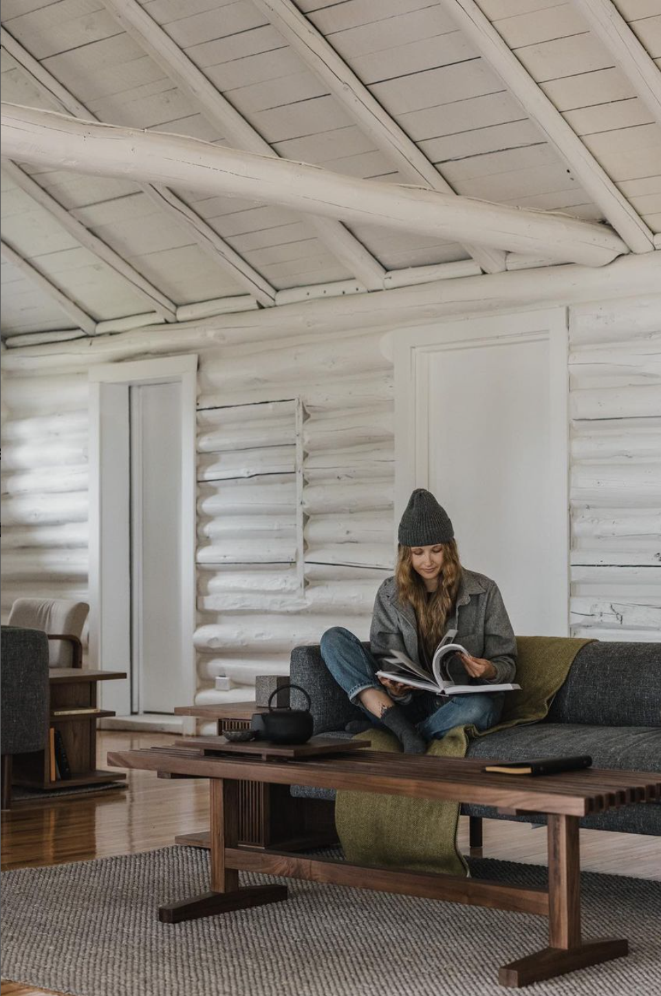 A woman sitting on a dark gray sofa, reading a magazine in a rustic, white-painted log cabin interior. She is wearing a gray beanie and a gray jacket, with a green blanket and black socks. There is a wooden coffee table with a black kettle and other small items in front of her. The room features white wooden walls, ceiling, and door frames, with light wood flooring.