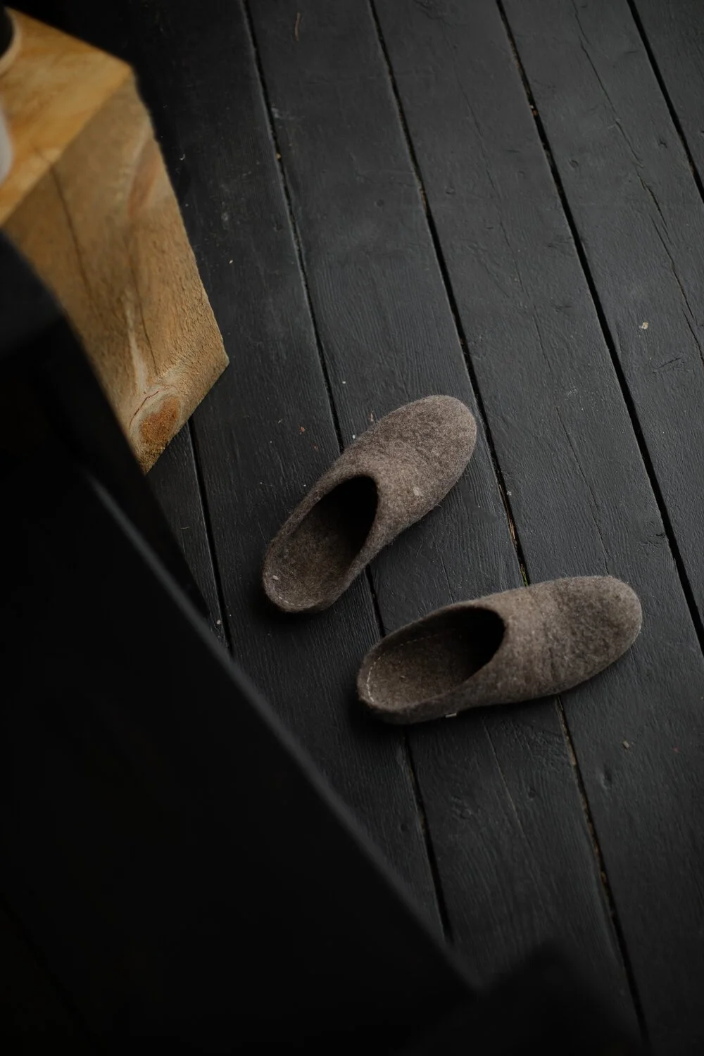 A pair of brown felt slippers on a dark wooden floor next to a wooden bench.