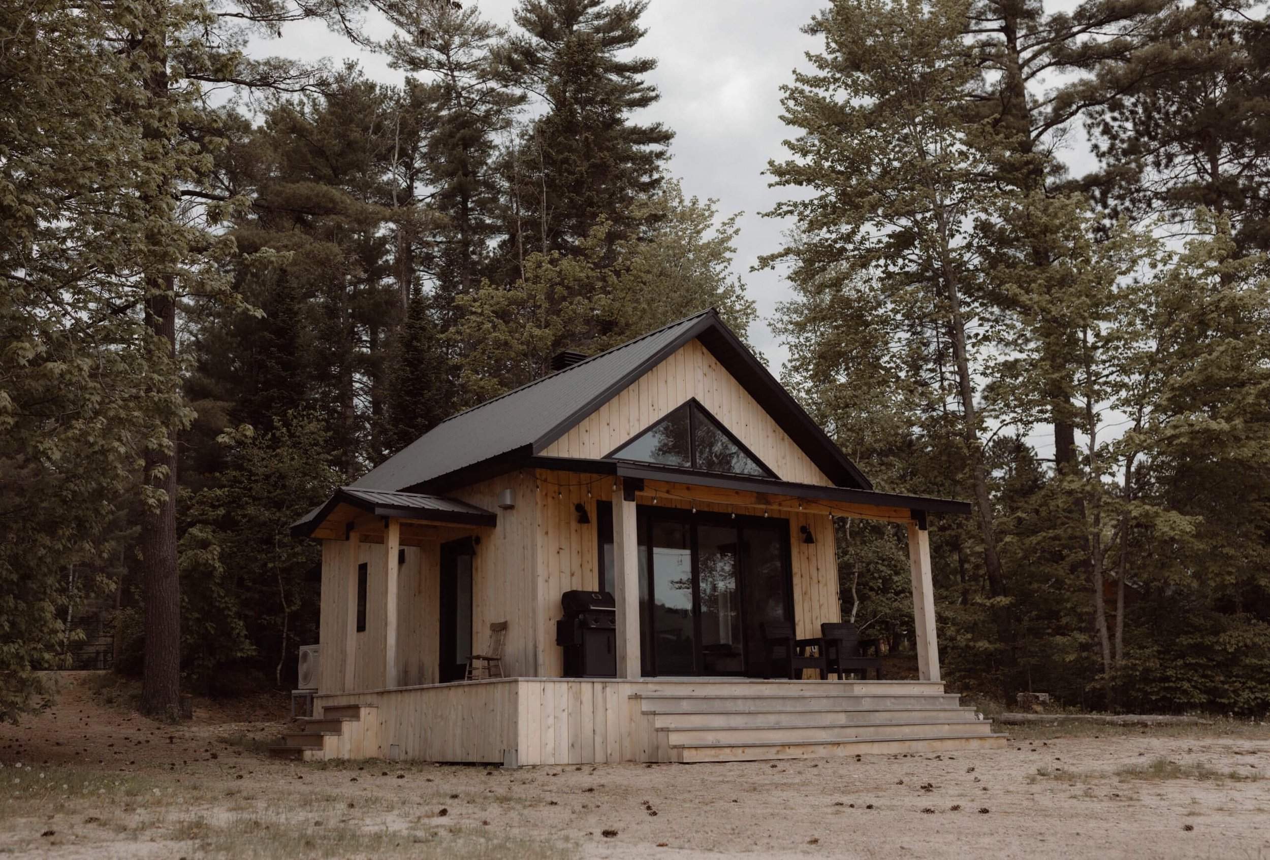 A small modern wooden house with a black metal roof, large glass doors, a porch with stairs, and outdoor furniture, surrounded by tall trees under a cloudy sky.