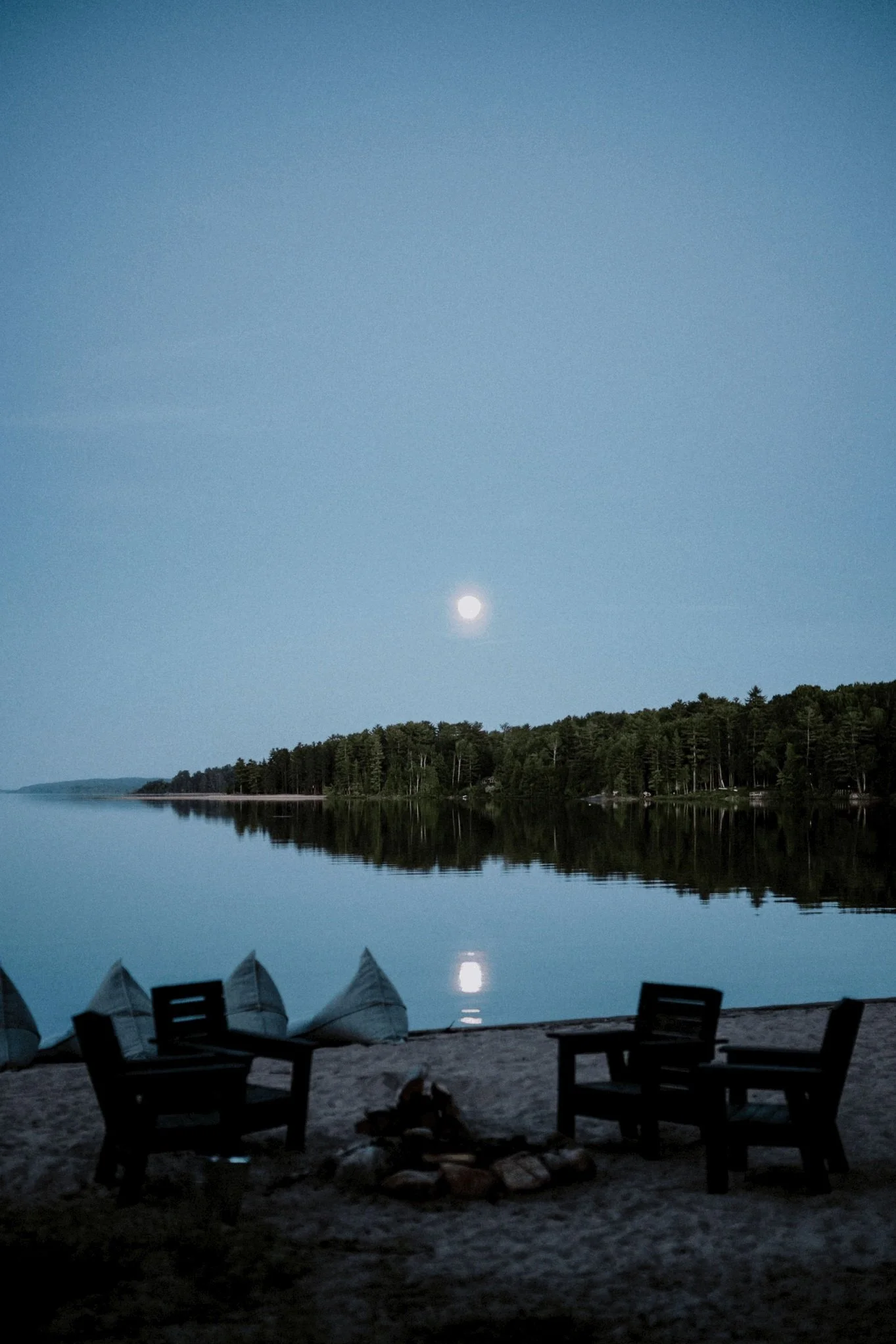 Nighttime scene by a lake with a bright moon above the horizon, the lake reflecting the moon, trees along the shoreline, and a beach area with chairs and a campfire.