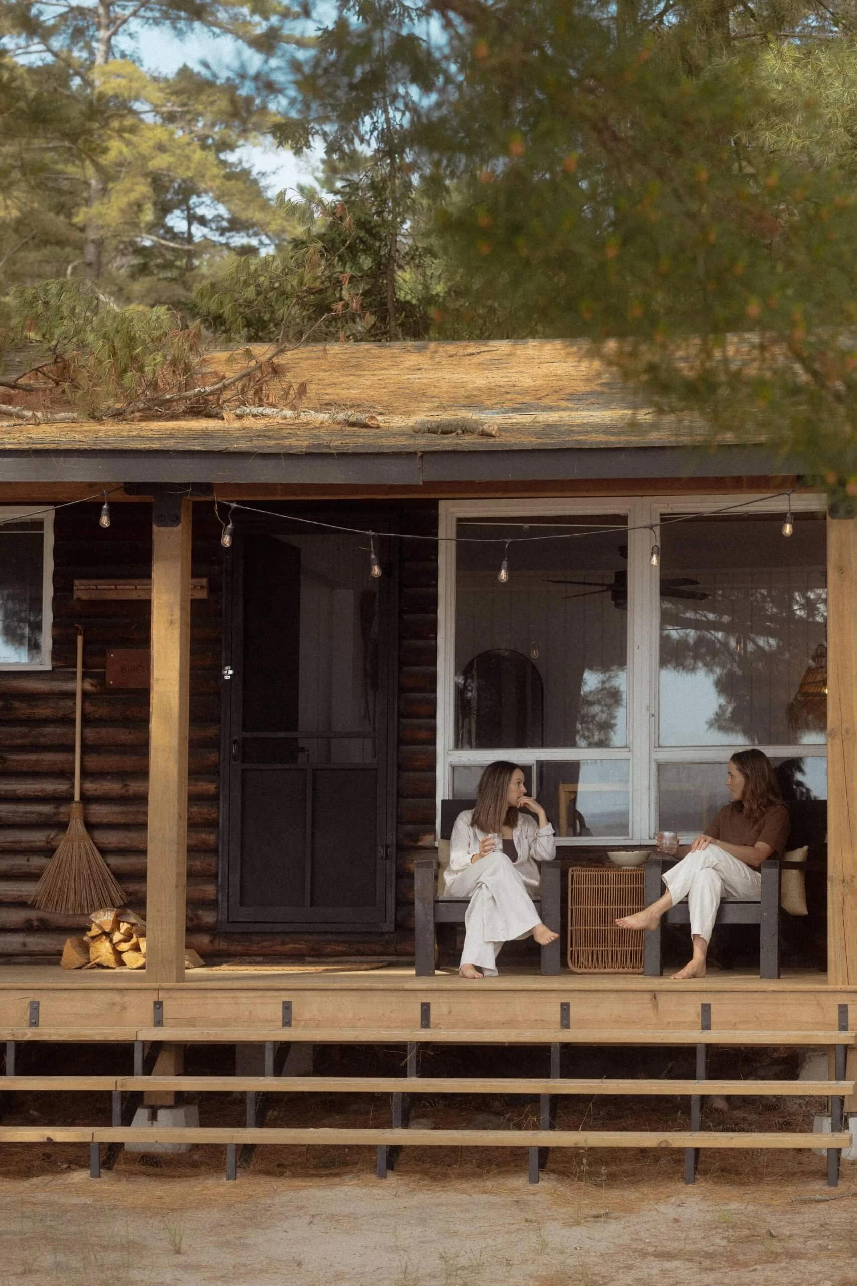 Two women sit and chat on a porch of a rustic cabin, surrounded by trees, with wood logs and a broom nearby.