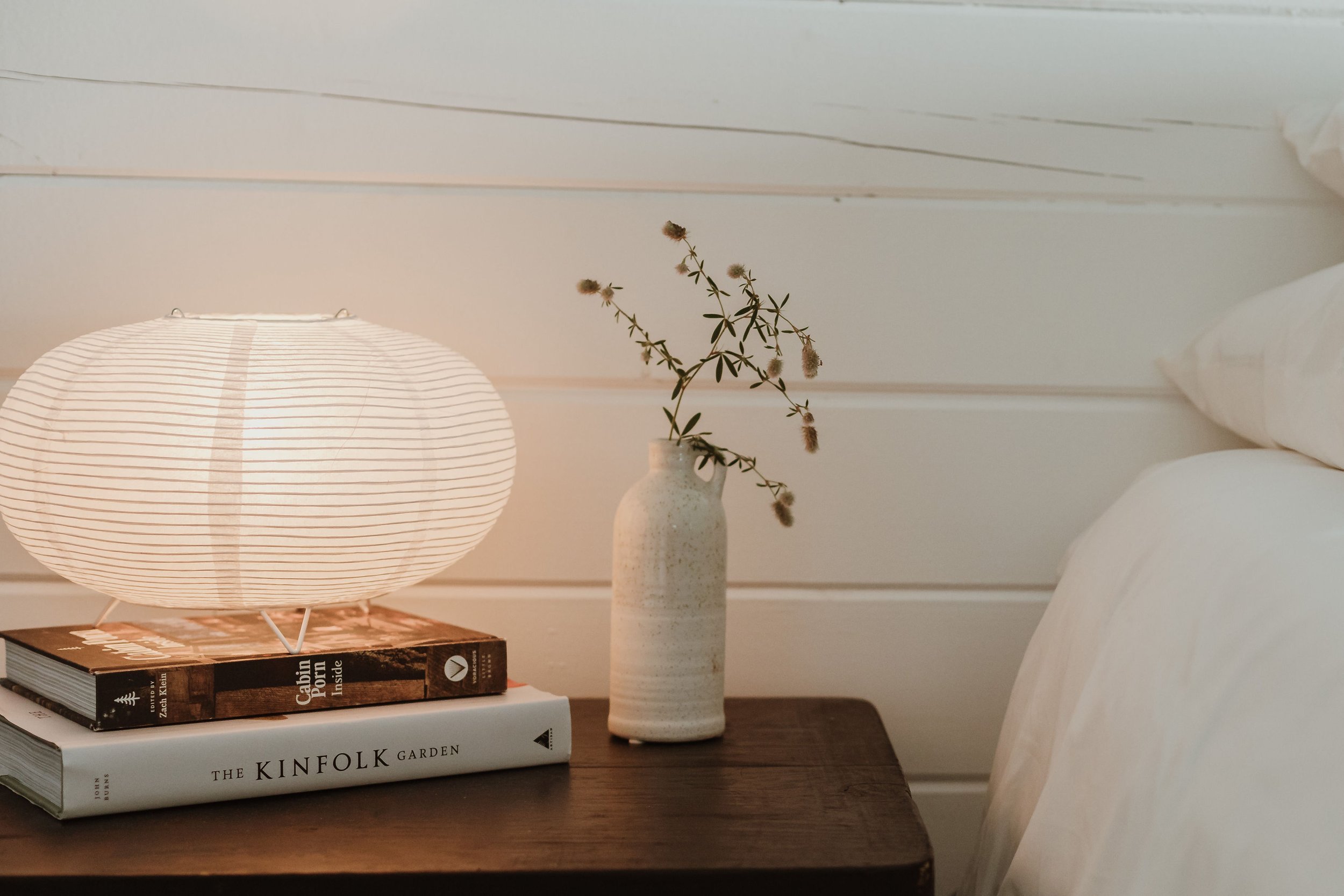 A wooden nightstand with a white lamp, a stack of two books titled 'Cabin Porn' and 'The Kinfolk Garden', and a ceramic vase with dried flowers. Behind the nightstand, there is a white wooden wall and a bed with white bedding.