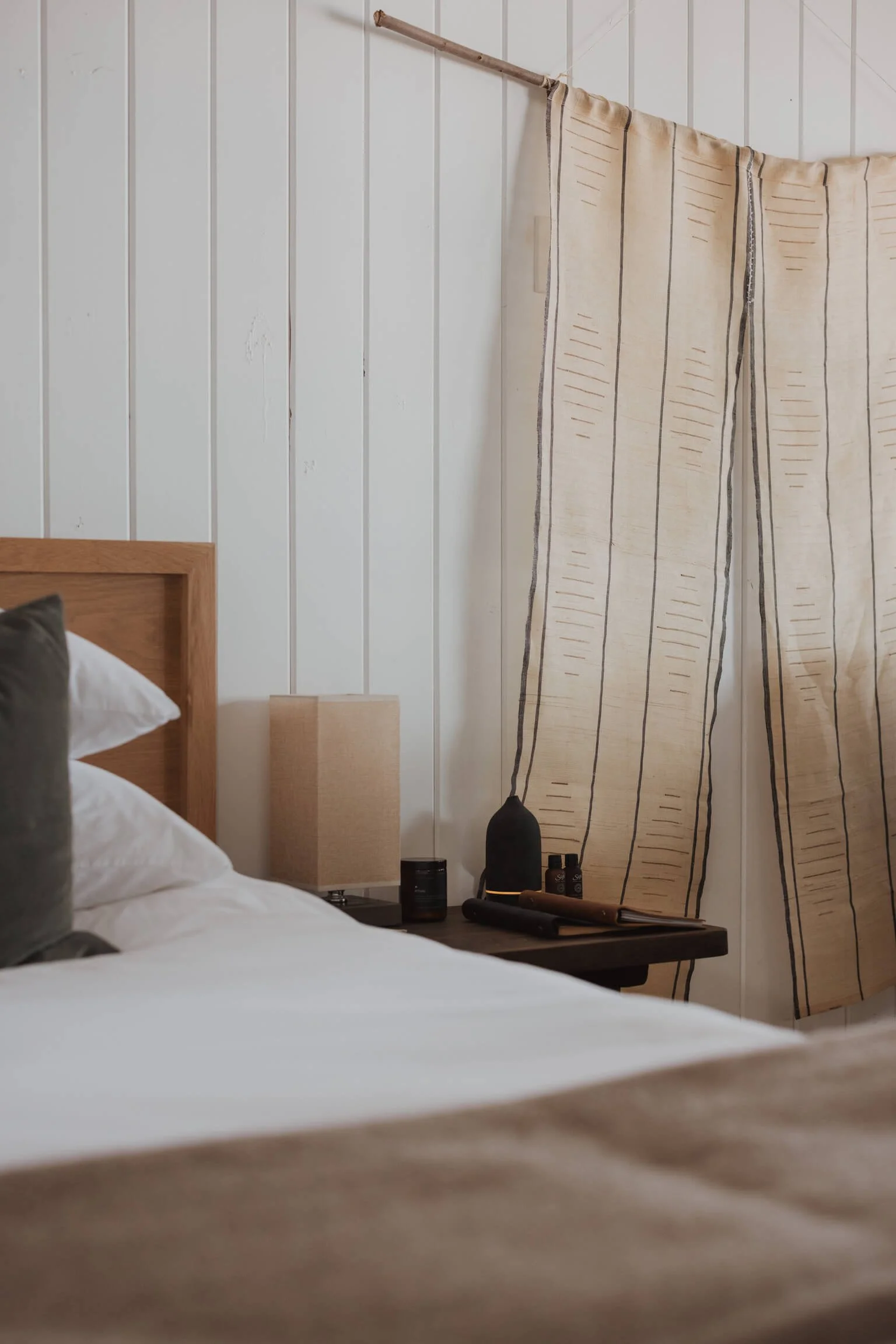 A cozy bedroom corner featuring a bed with white and gray pillows, a beige lamp on a dark wooden nightstand, and a cream-colored curtain with vertical black lines hanging on a wooden rod against white paneled walls.