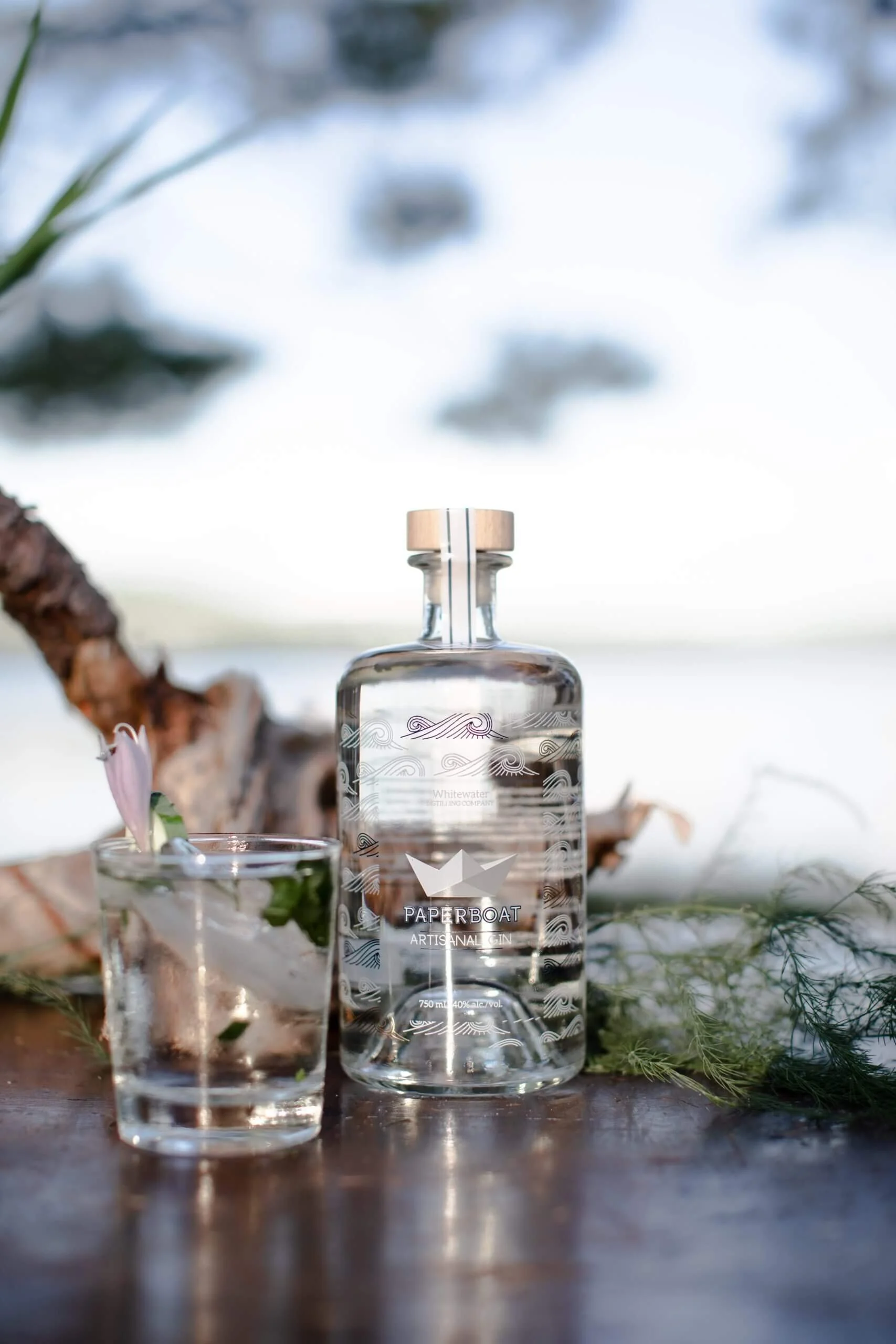 A bottle of Paper Boat gin and a glass with ice and mint leaves on a wooden surface outdoors with trees and sky in the background.
