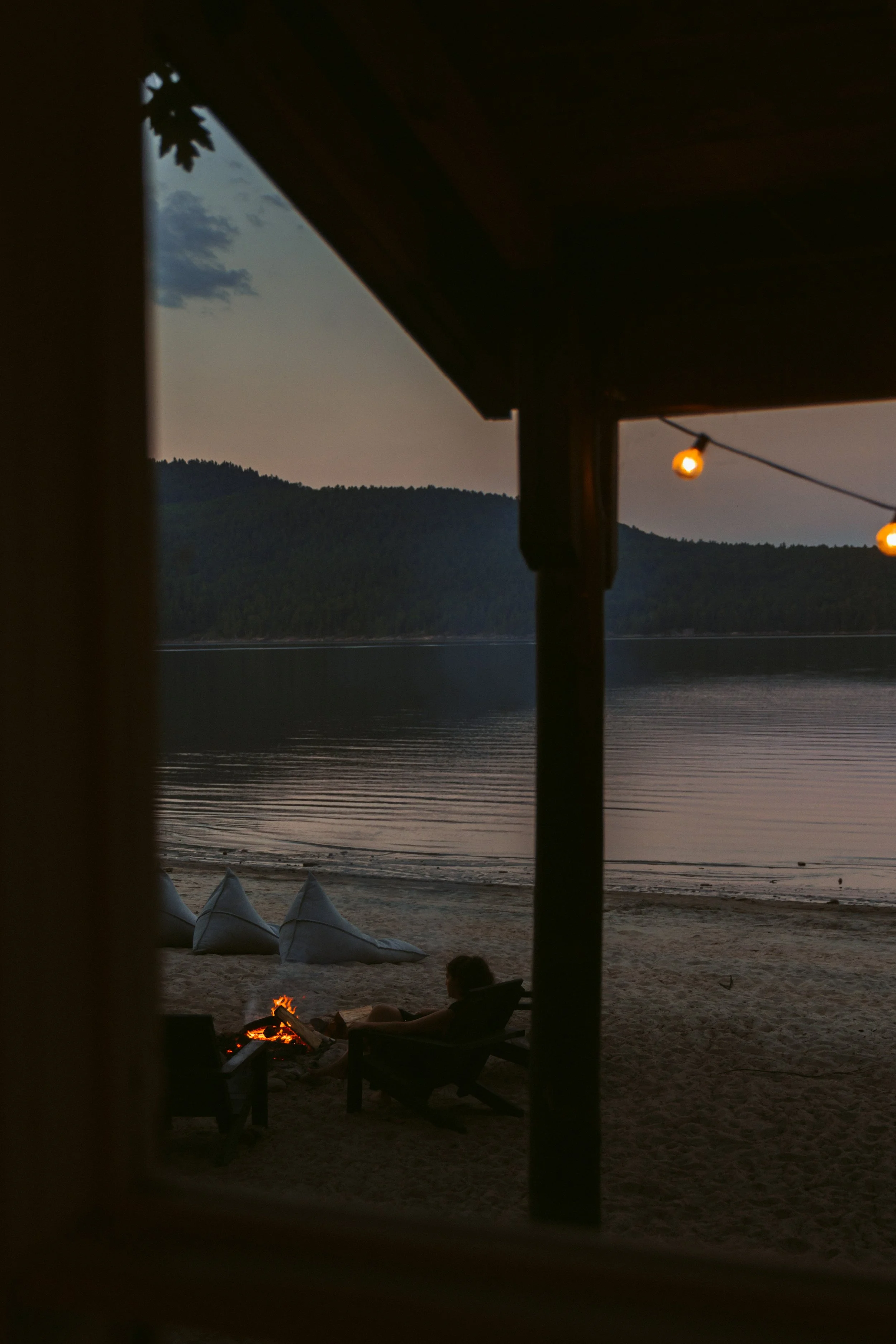 A person sitting by a campfire on a sandy beach at dusk, with a view of a calm lake and distant forested hills, seen through a window frame.