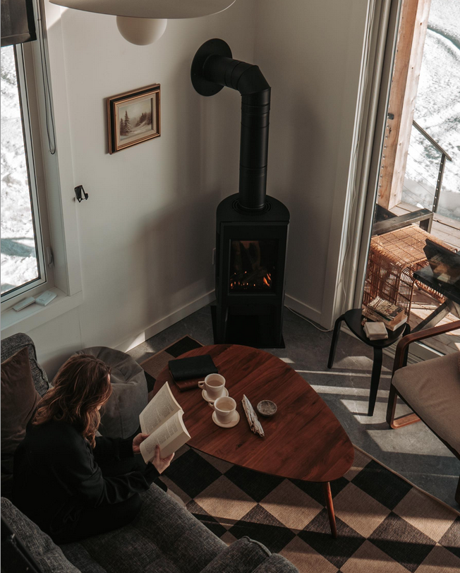 A woman sitting on a gray couch reading a book in a living room with a wood stove, a round wooden coffee table with cups and a book, a window showing snow outside, and a card chair near a glass sliding door.