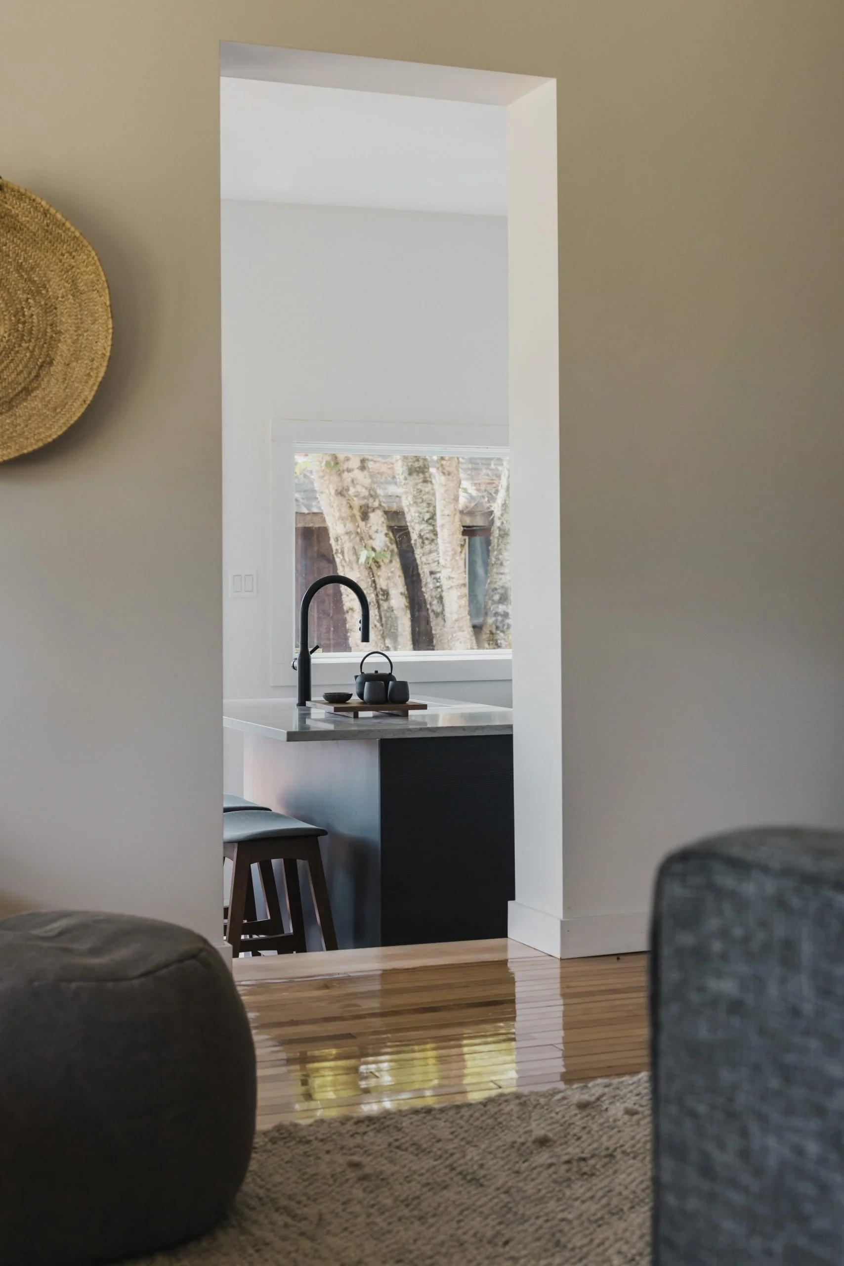 View through a doorway into a modern kitchen with a black sink and faucet, a window showing trees outside, and a marble countertop. The room is decorated in neutral colors with hardwood floors.