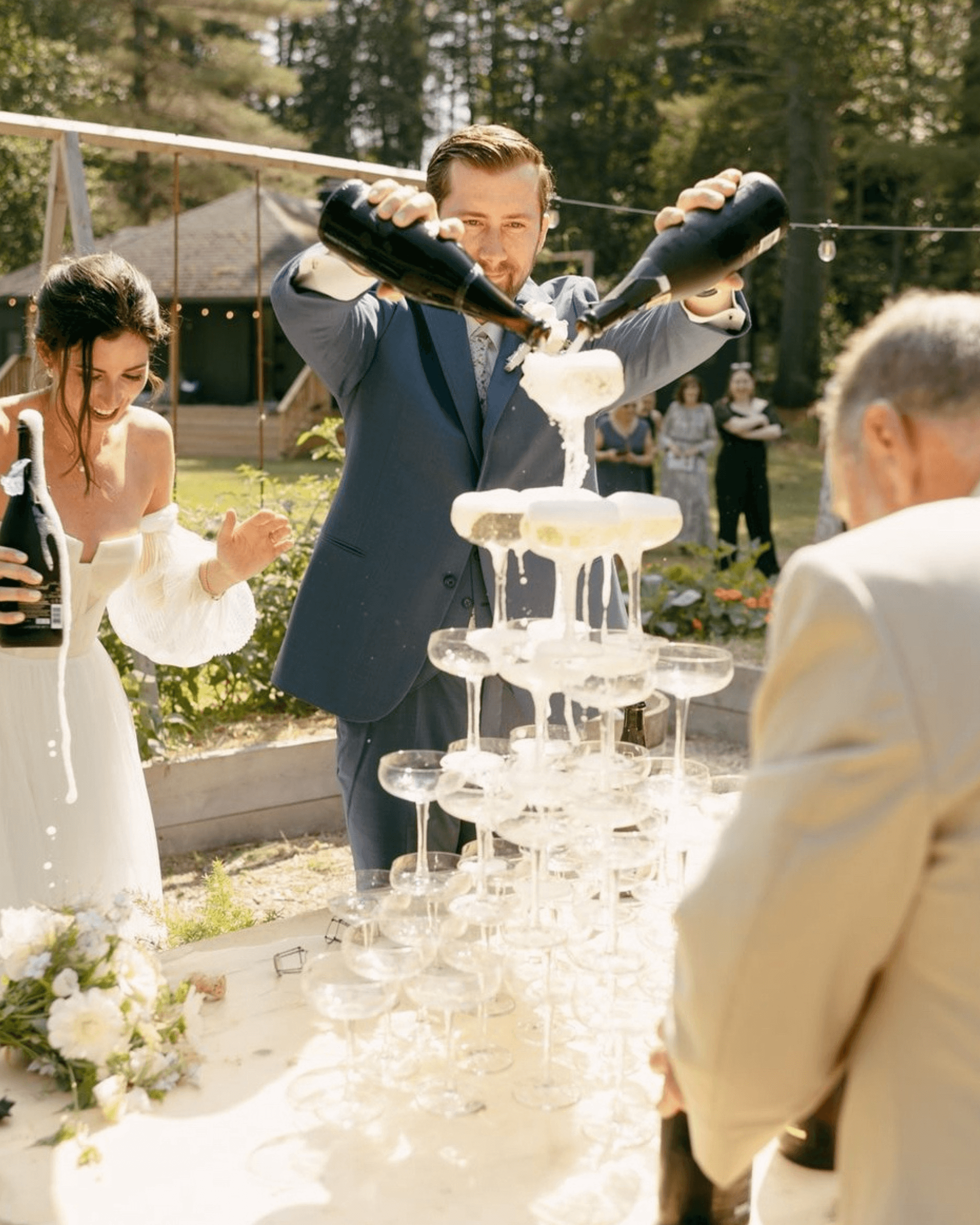 People celebrating outdoors as a man pours champagne into a pyramid of glasses during a wedding or party.