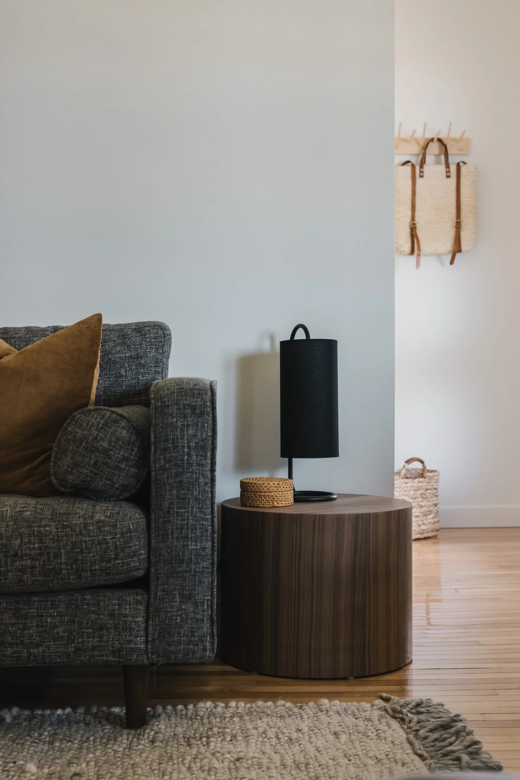 A cozy living room corner with a dark gray textured sofa, a brown pillow, a round wooden side table with a black lamp and a small woven basket, a light-colored wall with wall hooks and a beige basket on the hardwood floor.