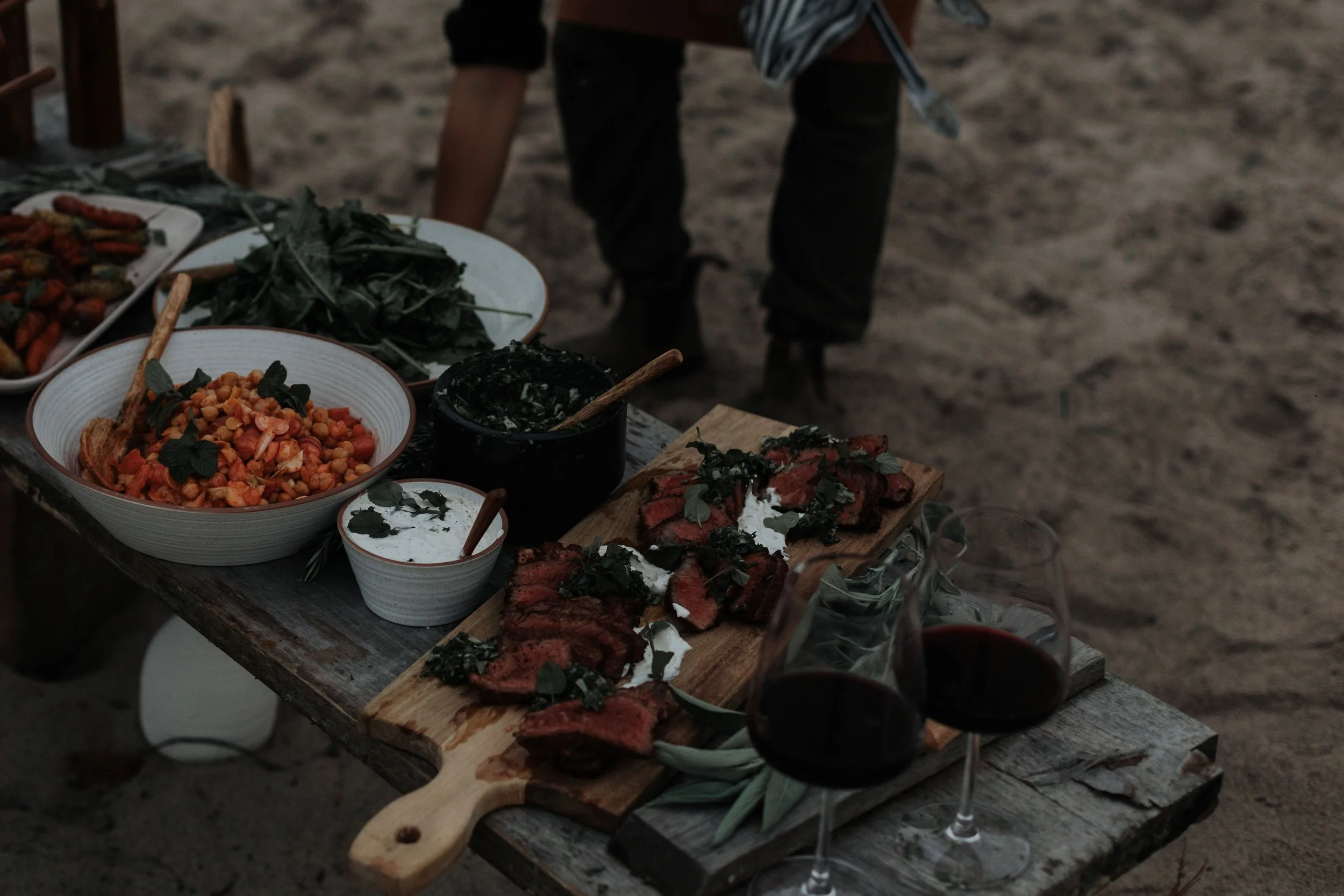 A rustic outdoor table set with various dishes including sliced steak, a chickpea salad, roasted vegetables, fresh herbs, and two glasses of red wine, with a sandy ground in the background.