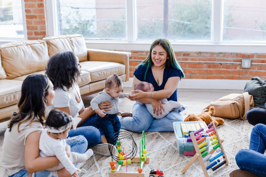 A group of women and children sitting on a carpeted floor in a brightly lit room with a brick wall and large windows. One woman is holding two babies, and a young girl is reaching out to touch a woman's face. Toys and personal belongings are scattered around.