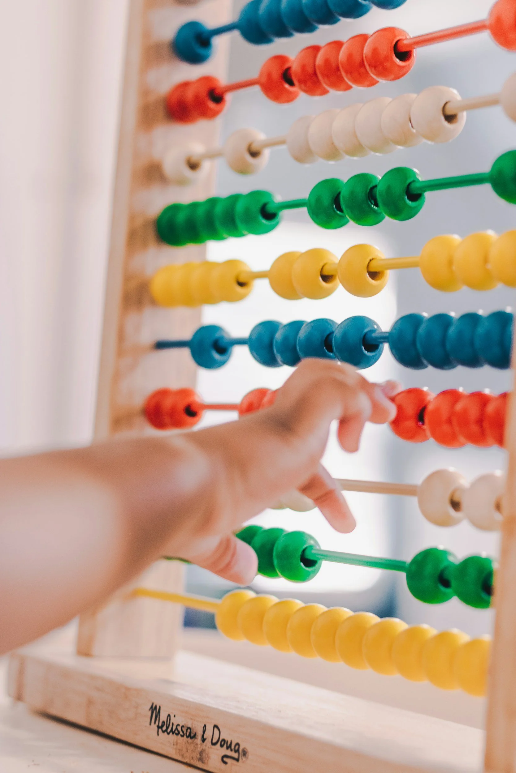 Close-up photo of a hand using an abacus with colorful beads in blue, orange, white, green, and yellow, on a wooden frame with a personalized engravement reading 'Melissa & Doug'.