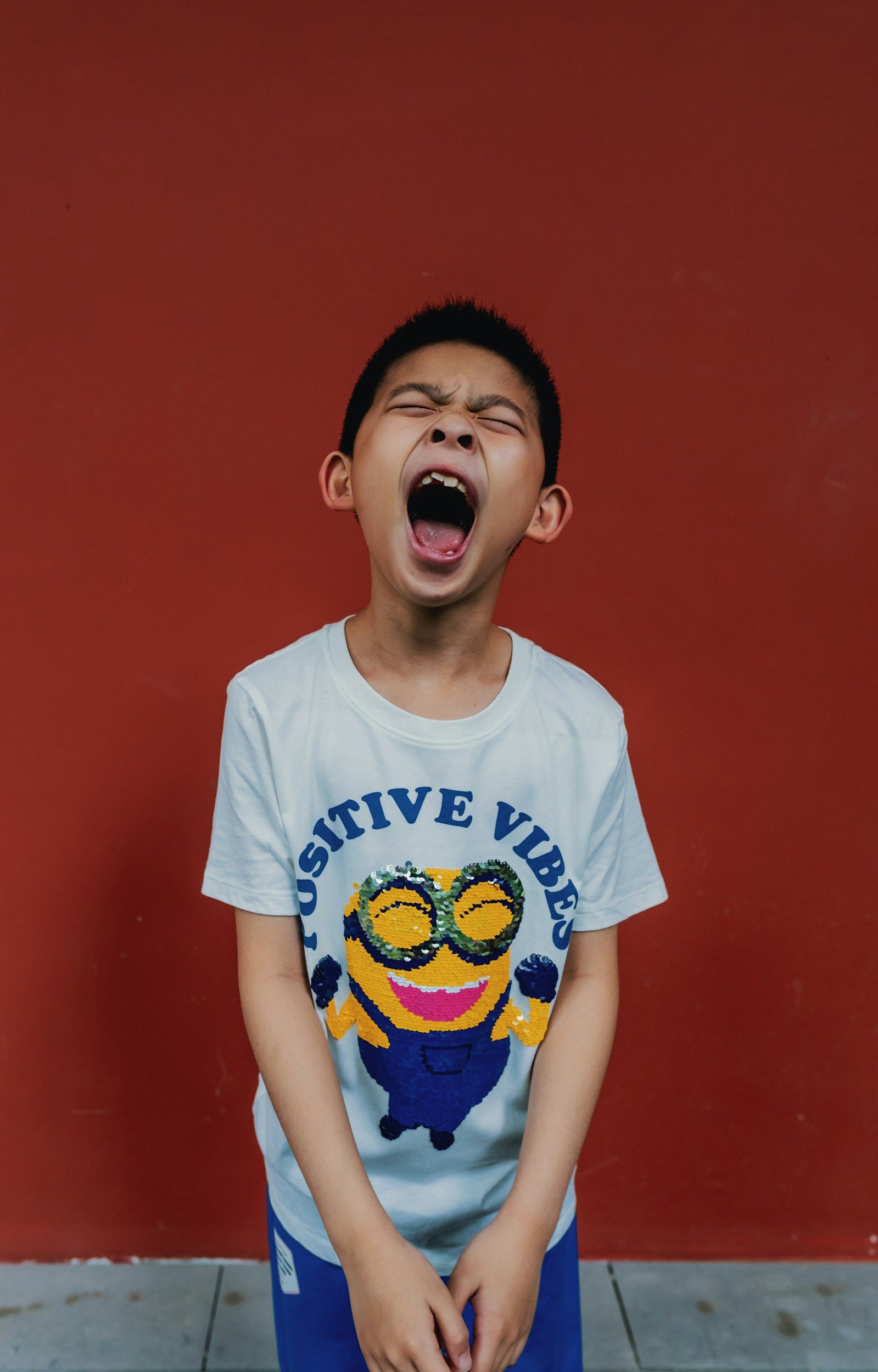 A young boy with short black hair, wearing a white T-shirt with a Minions graphic and the words 'Positive Vibes,' is standing against a red wall, yawning with his eyes closed and mouth wide open.