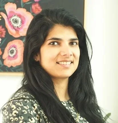 A woman with long black hair smiling, wearing a black and white patterned top, standing indoors in front of a floral artwork.