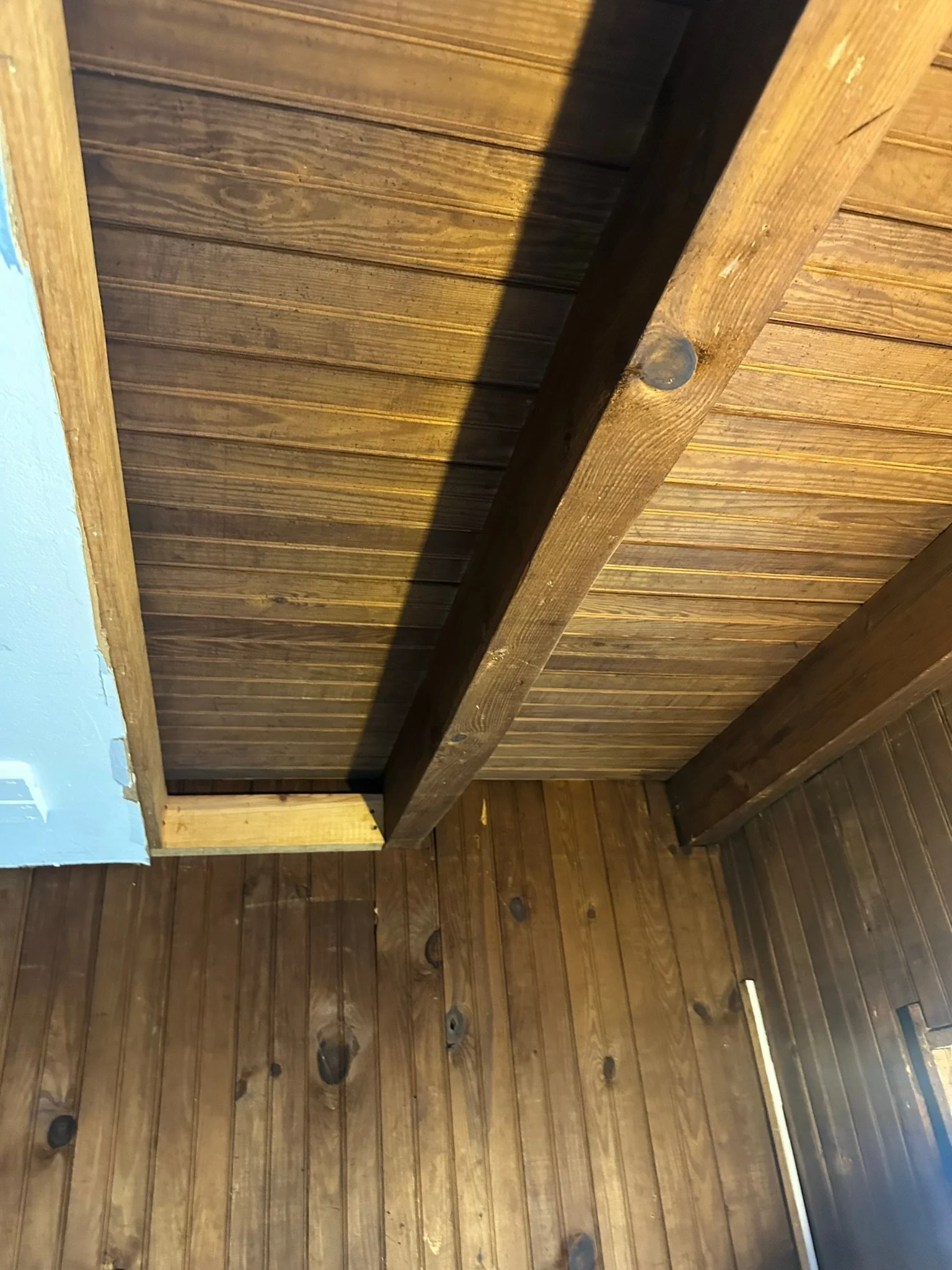 Wood-paneled ceiling and wall, with a shadow cast across the ceiling. Part of a floor with wooden planks and knots visible.