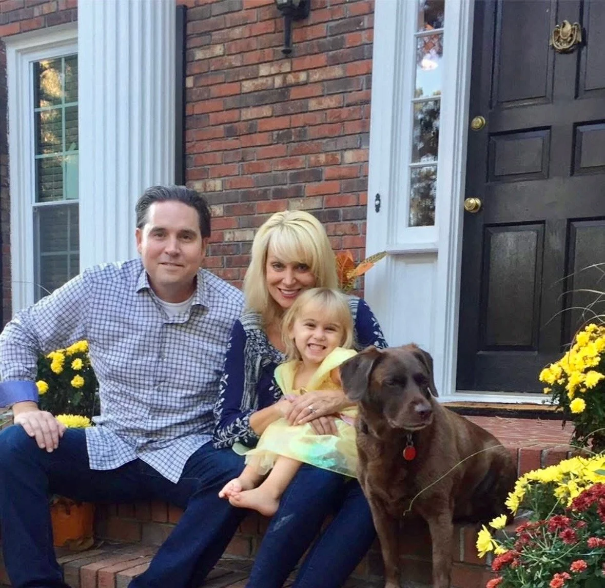 Family of three with a dog sitting on brick steps decorated with yellow and red flowers outside their home, next to a black door.
