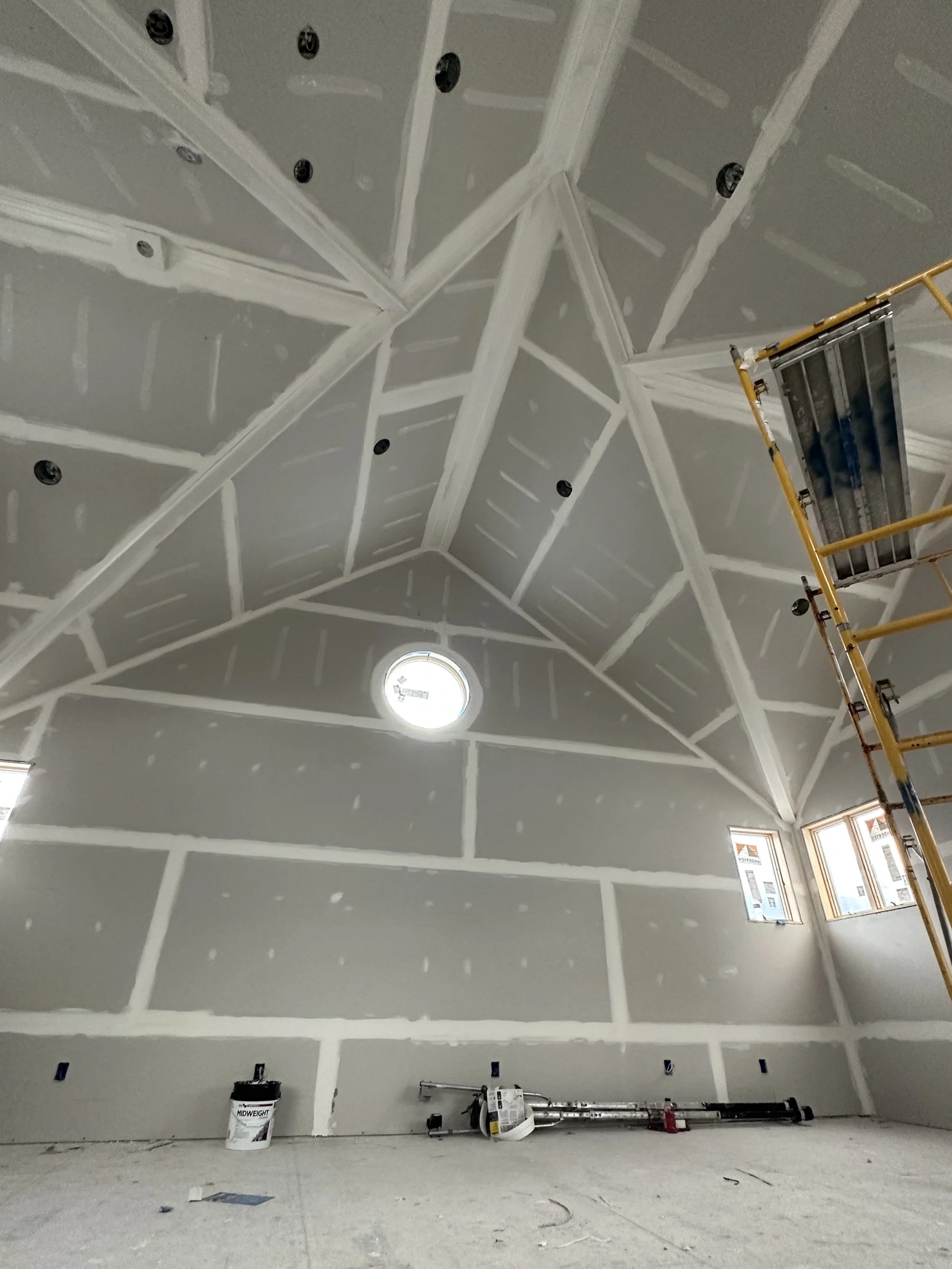 Interior of a room under construction with drywall installed on the walls and ceiling, with visible seams and joint compound. Construction equipment, including a scaffold, a bucket, and tools, are in the room.