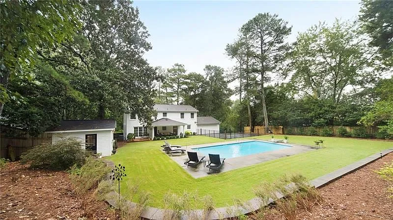 Backyard with a rectangular swimming pool, surrounded by a concrete deck with lounge chairs, lush green lawn, a small shed, tall trees in the background, and a white two-story house with a porch.