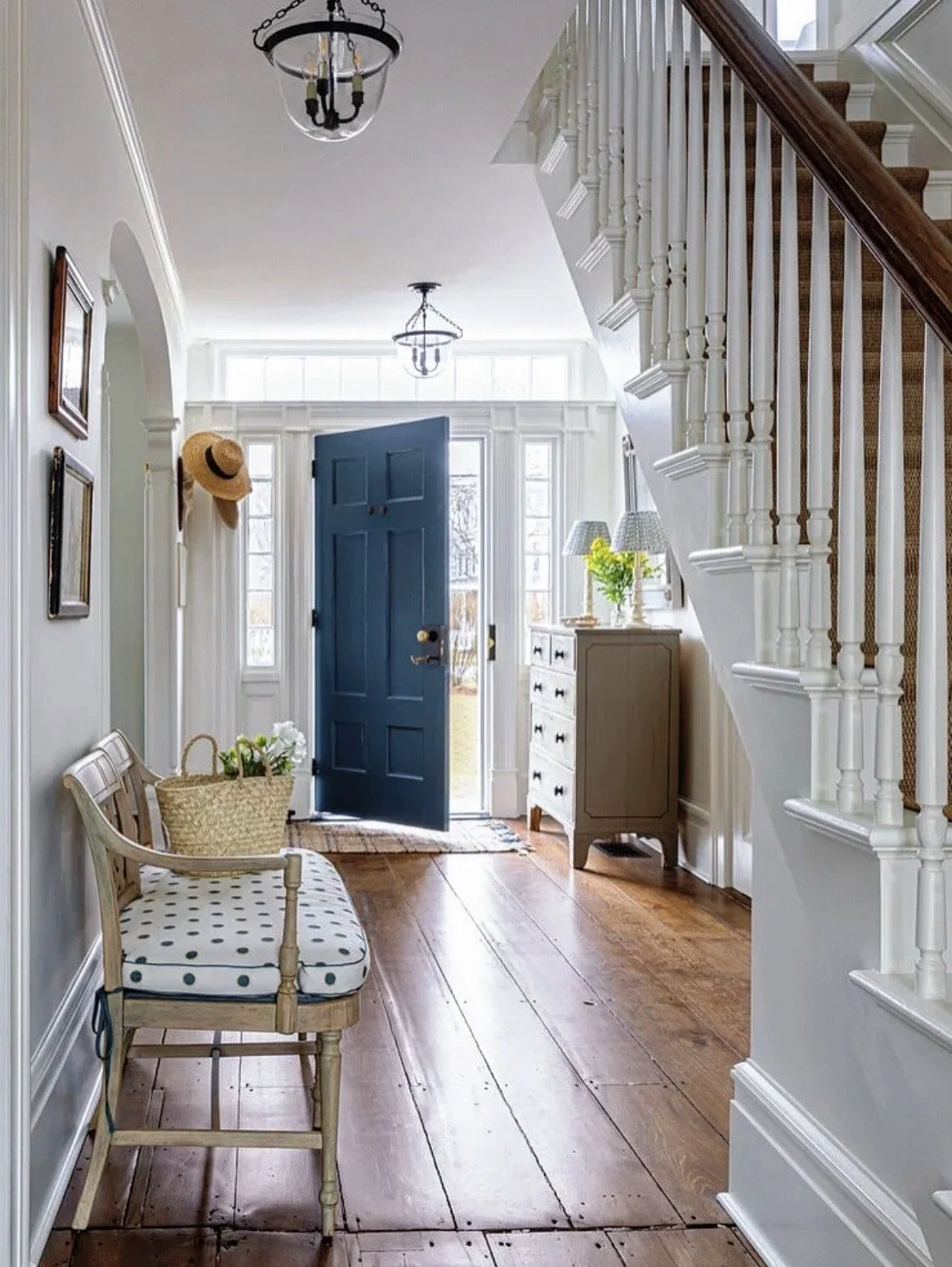 Entryway of a house with a blue front door open to the outside, white walls, wooden floor, a bench with polka-dot cushion, a basket with flowers, a small gray dresser, and a staircase on the right side.