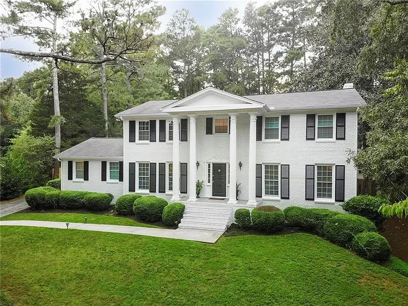 A large, two-story white house with black shutters, surrounded by well-manicured bushes and green lawn, set against a wooded background.