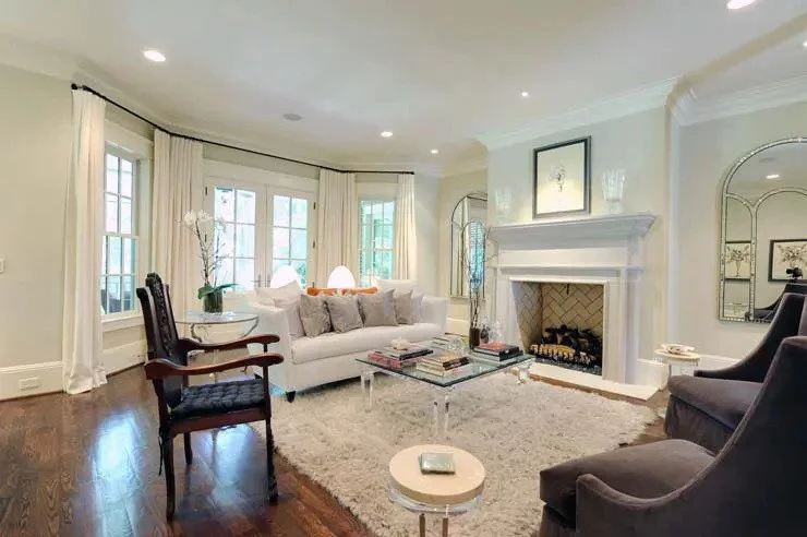 Bright living room with white walls, large windows with white curtains, white sofa, dark armchairs, glass coffee table, fireplace, decorative mirror, and framed artwork.
