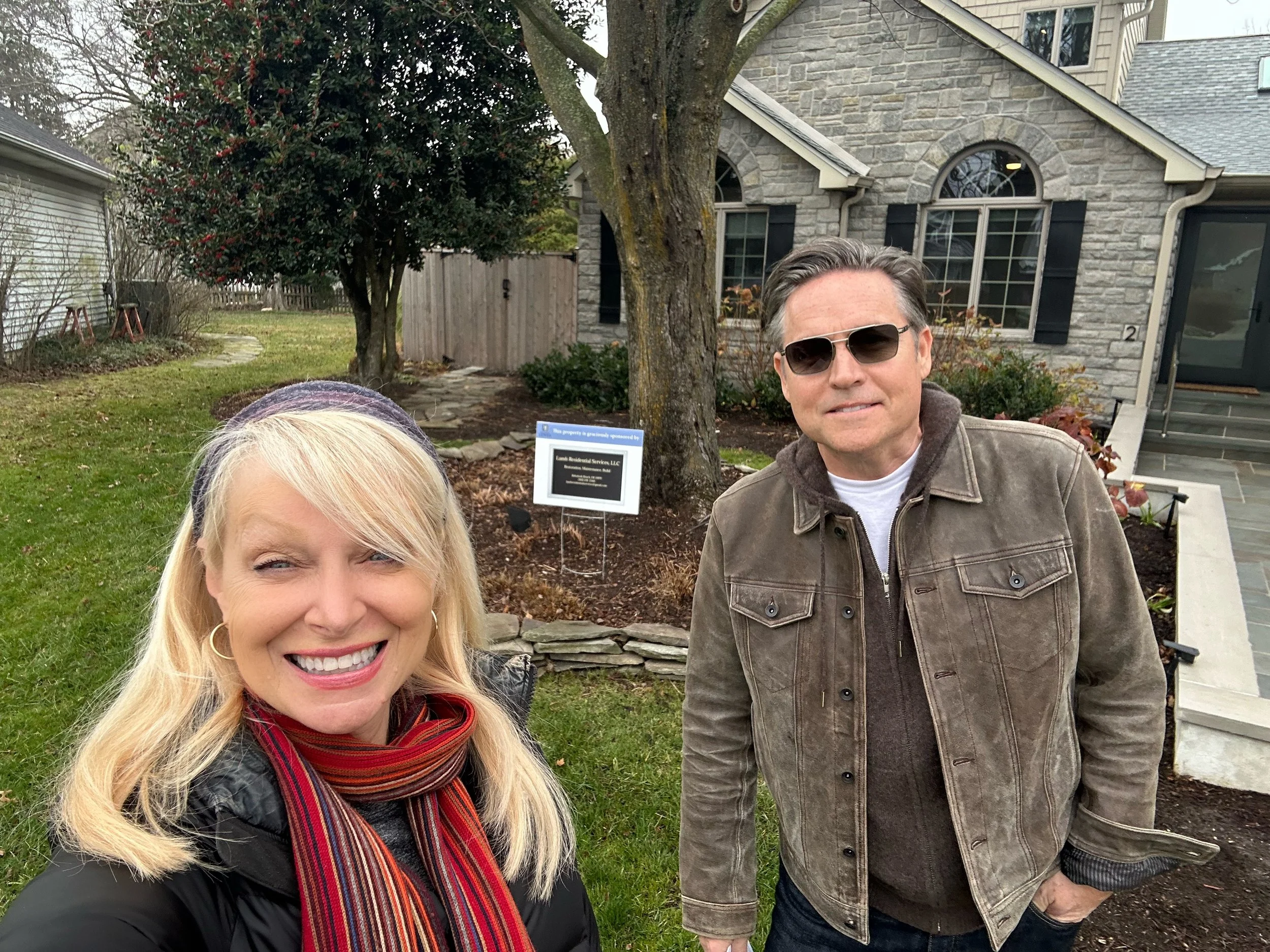 Smiling woman with long blonde hair and a scarf taking a selfie with a man wearing sunglasses and a brown jacket in front of a house with a garden.