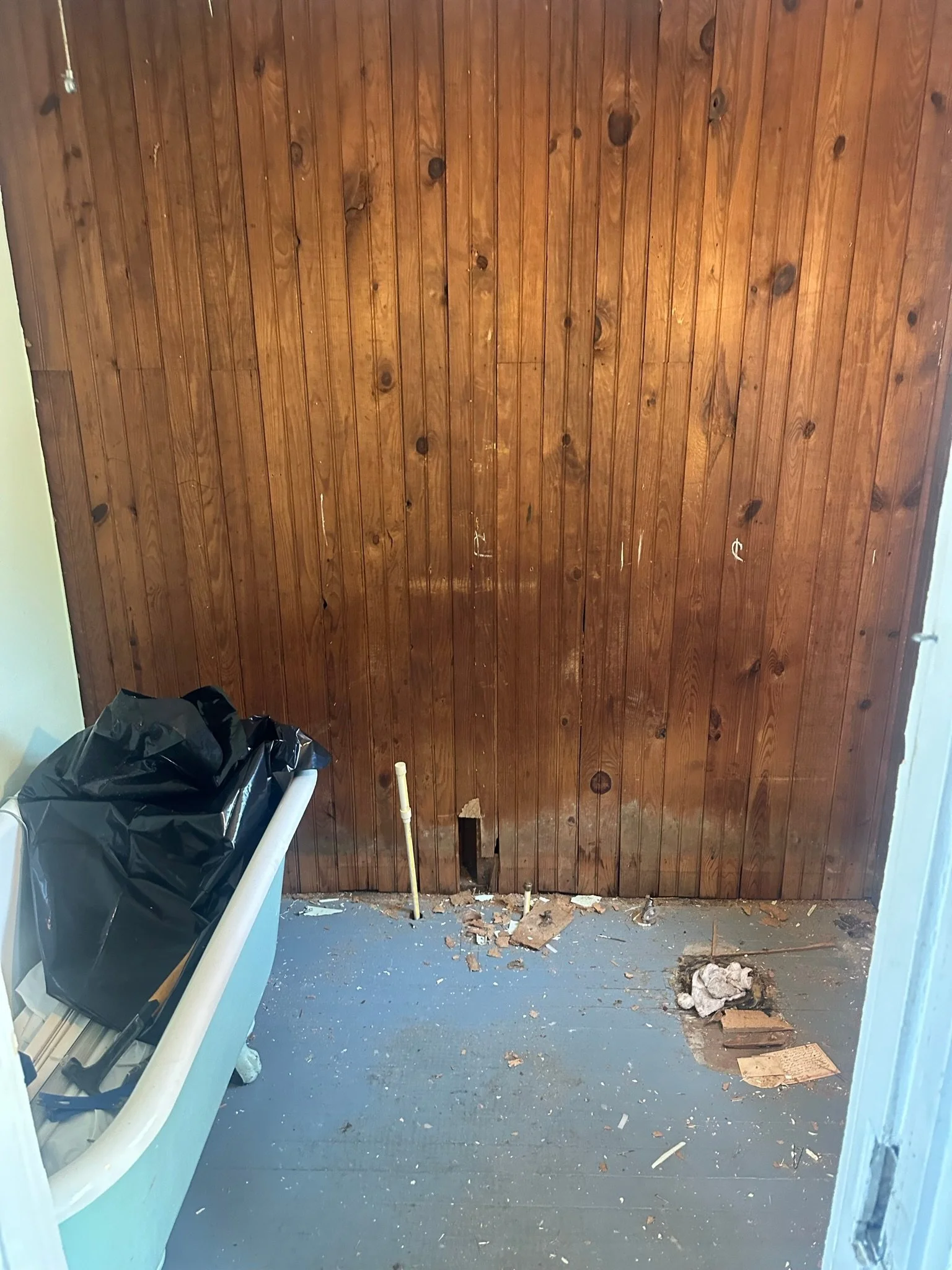 A room with a wooden wall, a hole at the bottom of the wall with bricks and debris scattered on the floor, a yellow pipe to the left of the hole, a plastic trash bag on top of a light blue container, and a white pole leaning against the wall.