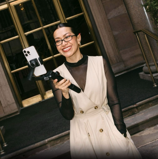 Woman smiling wearing glasses and cream jumpsuit holding phone on grip for capturing content.
