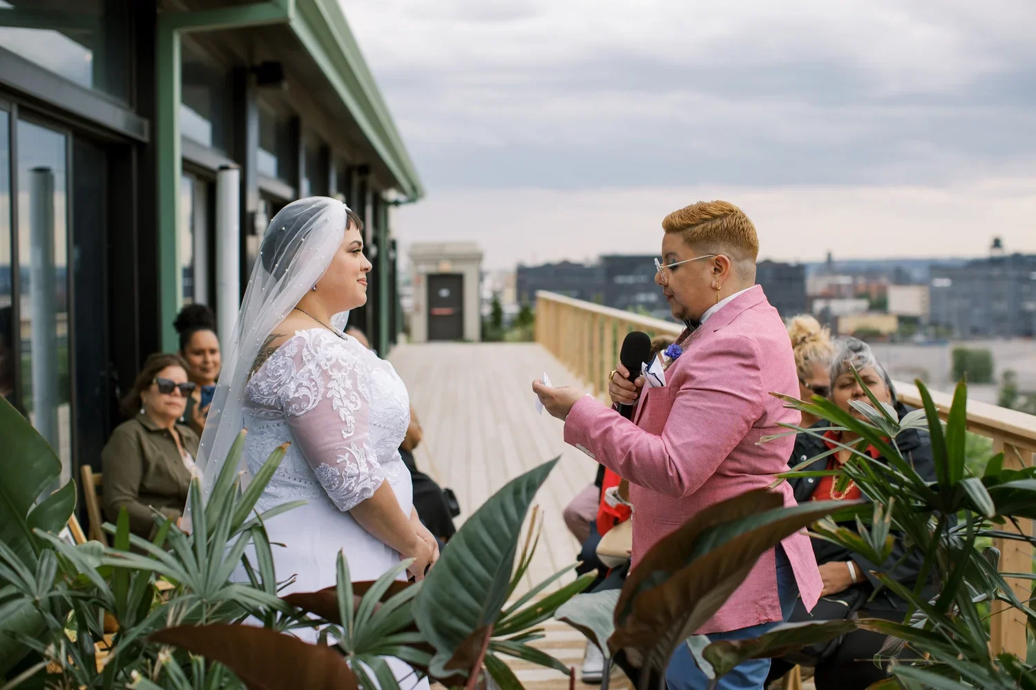 A queer couple in a white wedding dress and a pink blazer read their vows on an outdoor balcony with guests sitting behind them and city skyline in the background.
