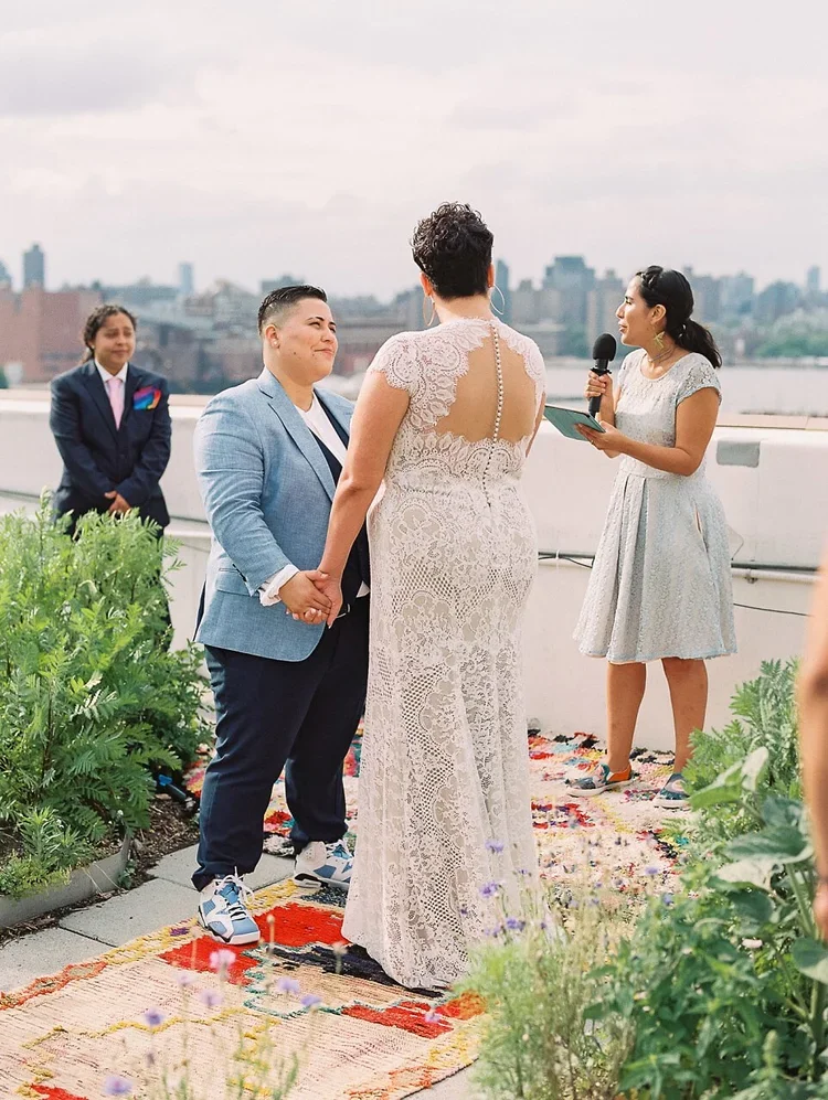 exchanges vows on a rooftop with a city skyline in the background, during a wedding ceremony. One partner wears a white lace dress, and the other wears a light blue blazer and dark pants. An officiant stands to the right, holding a microphone and reading from a tablet