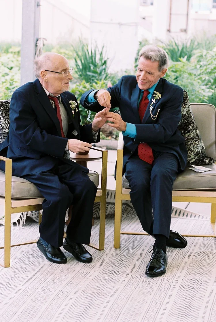 Gay wedding of two older grooms in suits exchanging wedding rings in an indoor garden setting.