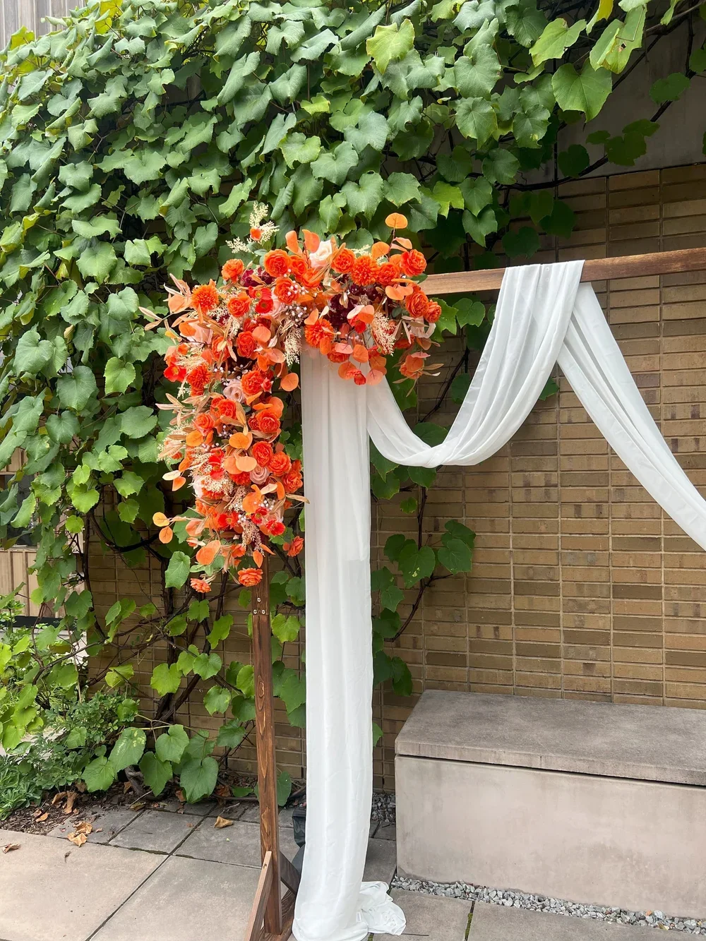 A decorative wedding arch decorated with orange and peach flowers and white drapery, standing outdoors against a brick wall and green leafy vines.