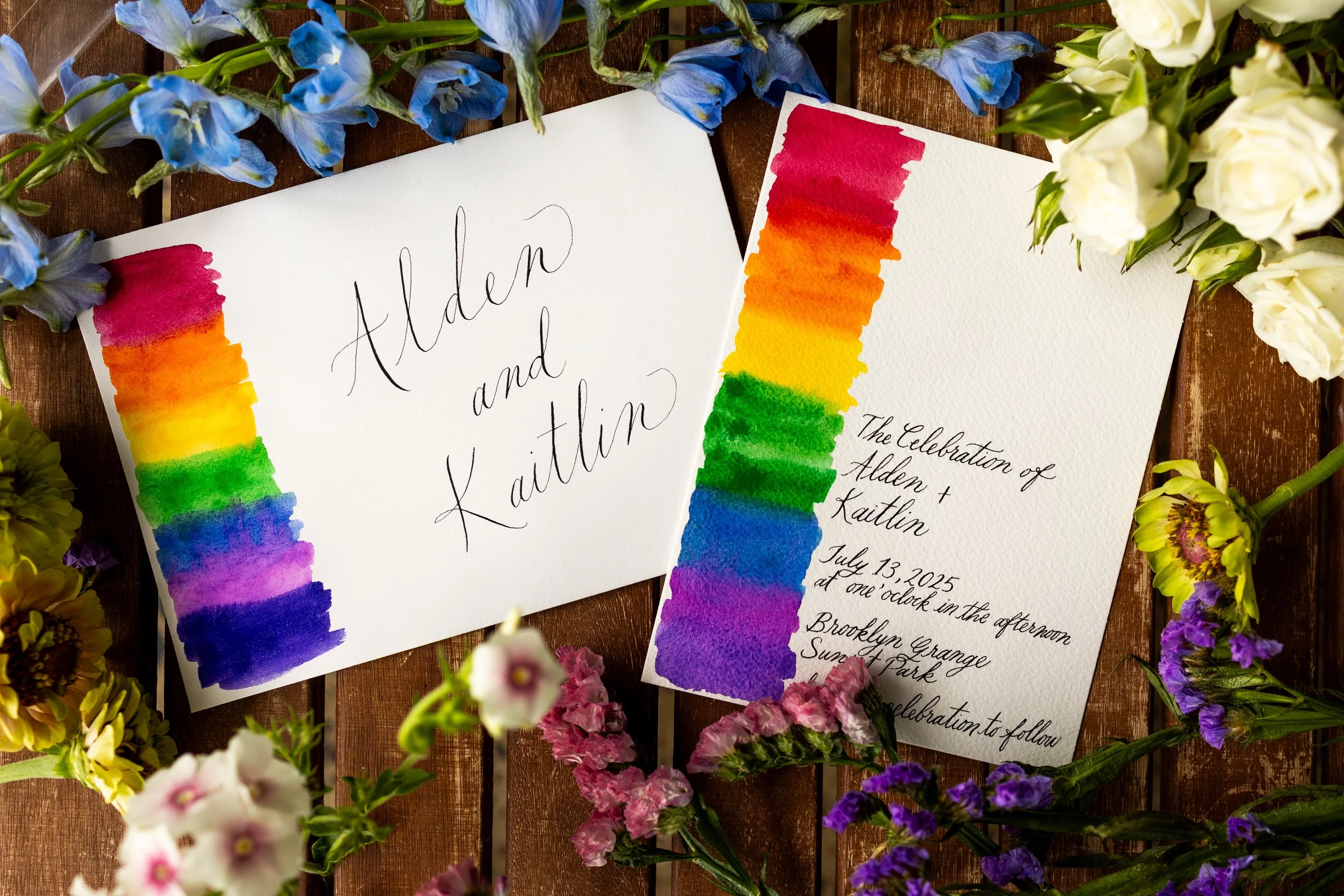 Two invitation cards with rainbow-colored borders, surrounded by various colorful flowers on a wooden surface. One card has handwritten text, "Alden and Kaitlin," and the other provides event details, including date, time, and location.