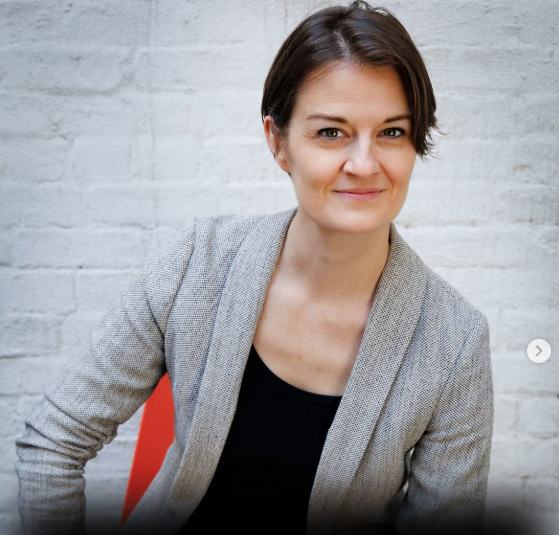 Portrait of a woman with short brown hair, wearing a gray blazer and a black top, sitting against a white brick wall.