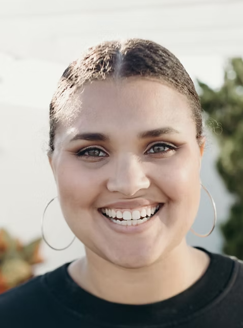 A smiling woman with hoop earrings, light makeup, and her brown hair pulled back, standing outdoors with a blurred green and white background.