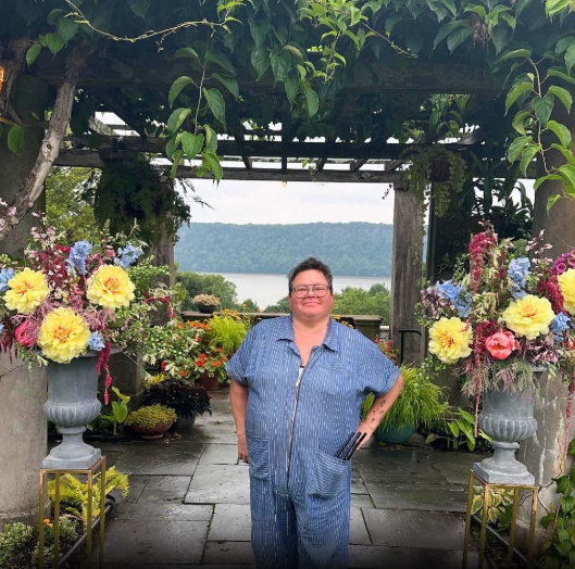 Person standing outdoors in a garden with colorful flower arrangements, a lake in the background, and a wooden pergola covered with green foliage.