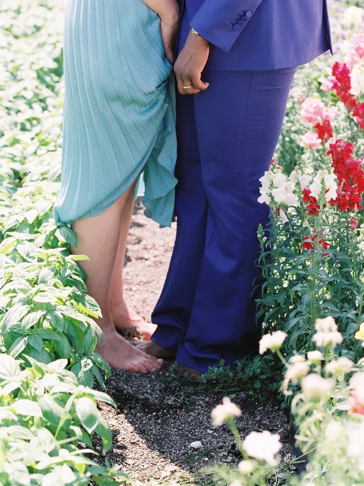 A queer couple holding hands, standing barefoot on a garden path surrounded by colorful flowers.