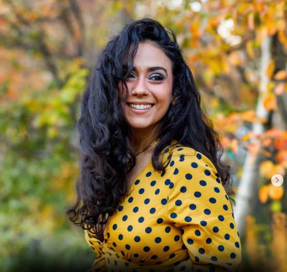 A young woman with long curly dark hair smiling outdoors in front of autumn trees, wearing a yellow dress with black polka dots.