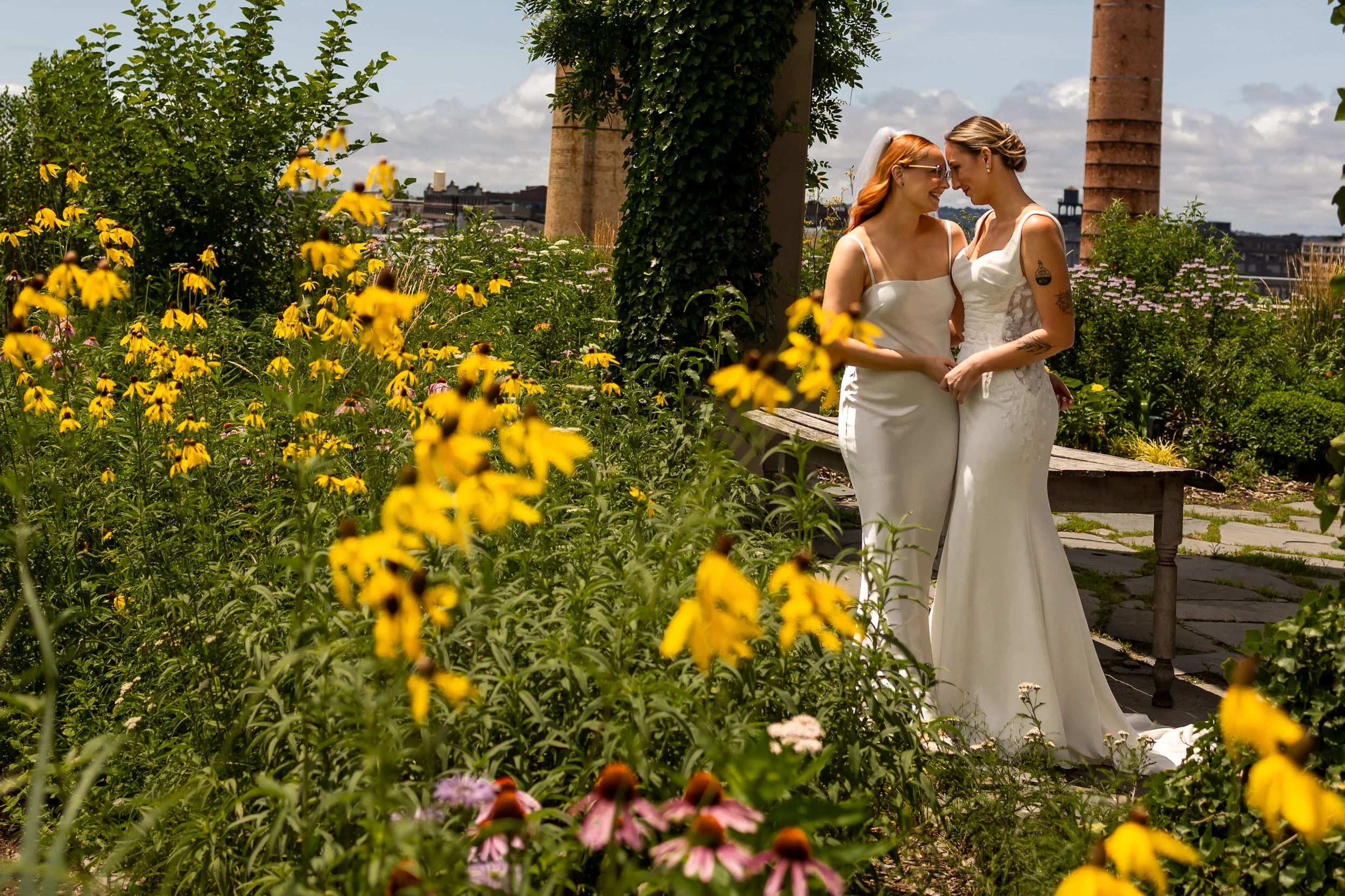 Queer wedding of two brides in white wedding dresses standing close together in a lush garden with yellow flowers, smiling and touching foreheads, with city buildings and chimneys in the background.