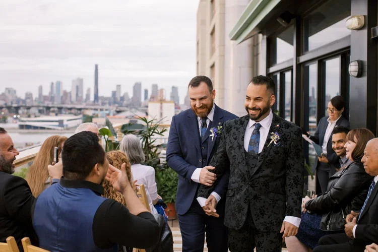 Gay wedding of two grooms in suits, one in a black floral suit, walking arm-in-arm at an outdoor event, surrounded by seated guests and city skyline in the background.