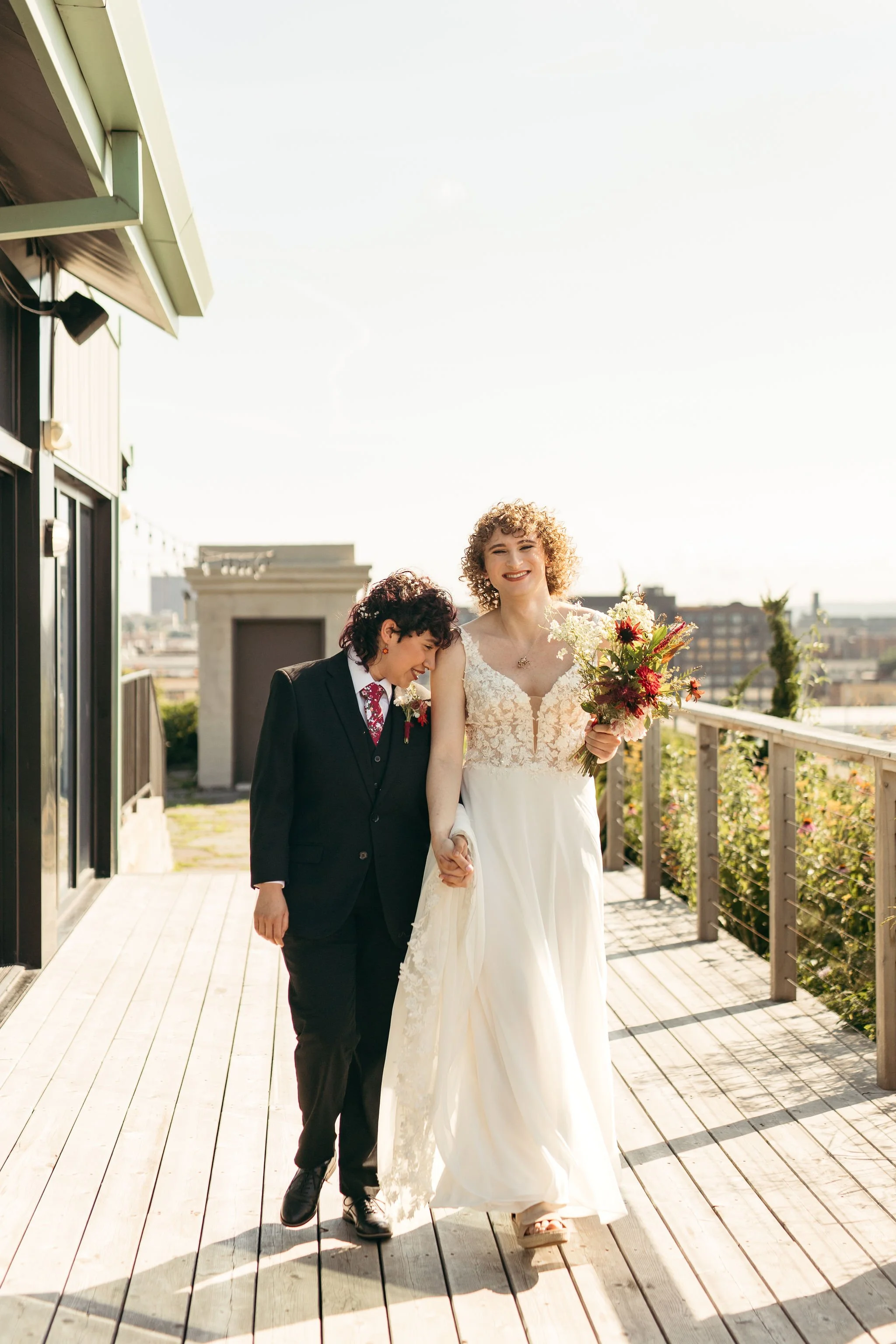 Two women walking hand in hand on a sunny outdoor balcony, one wearing a white wedding dress and holding a bouquet, the other dressed in a black suit with a red and white floral tie.