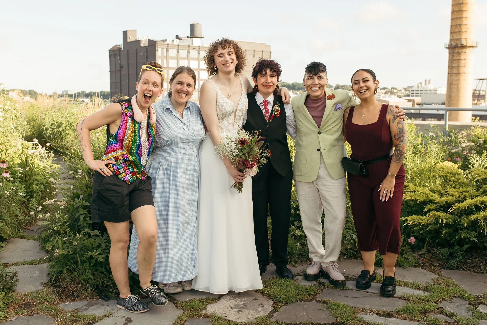 Group of seven people standing outdoors, smiling, with greenery and city buildings in the background, celebrating a wedding.