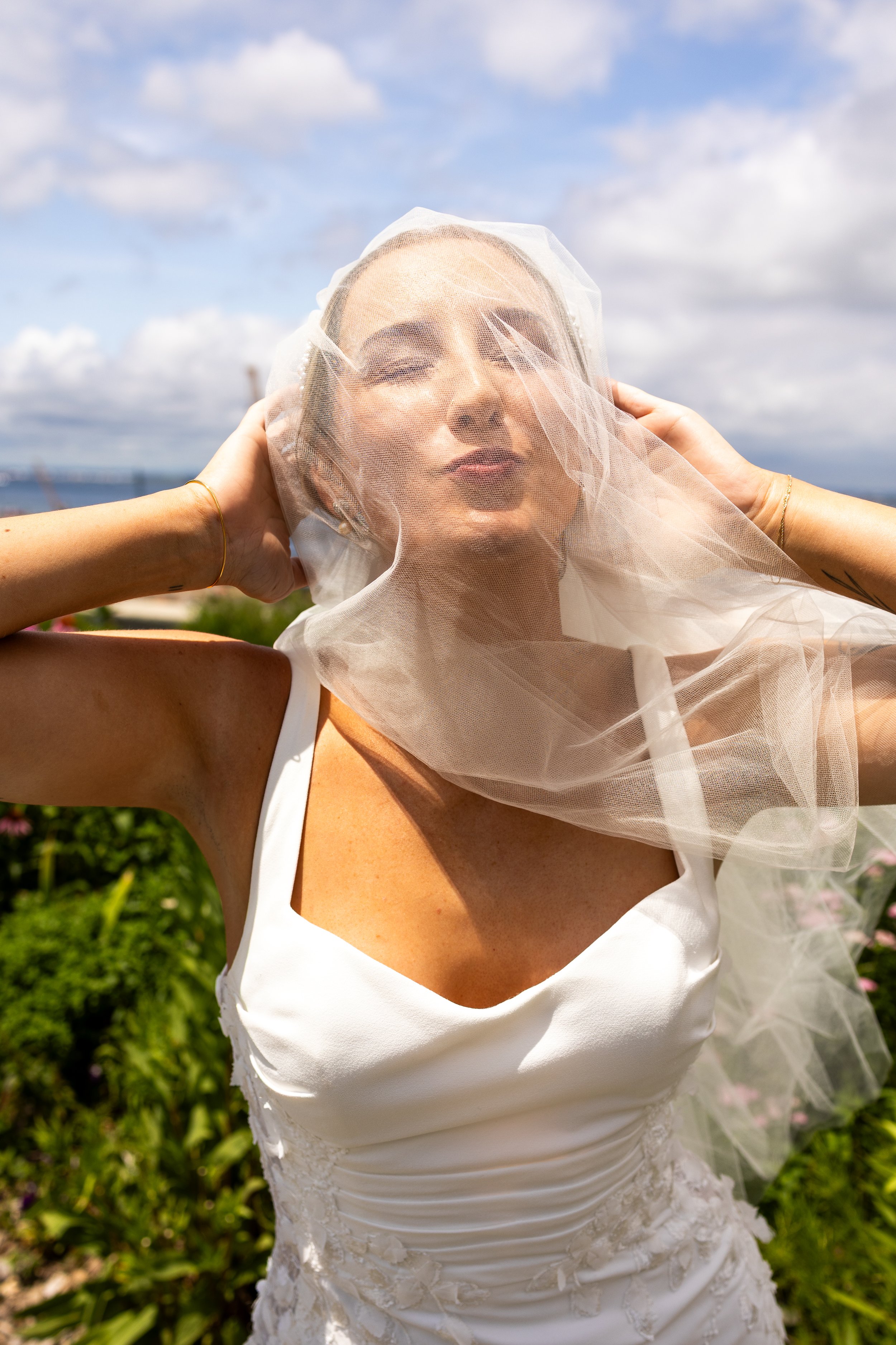 A queer bride in a white dress and veil outdoors, holding the veil over her face, with a cloudy sky and greenery in the background.