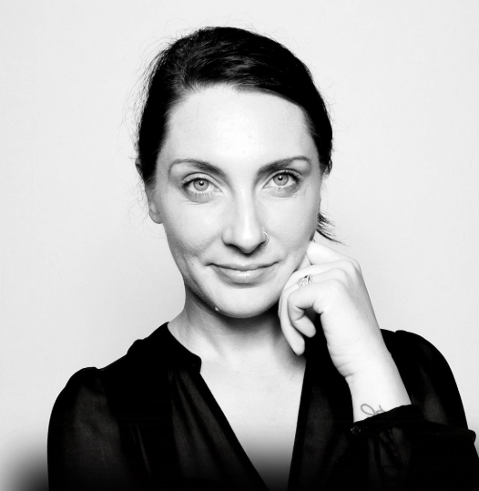 Black and white portrait of a woman with dark hair, fair skin, and light eyes, touching her face with her right hand, wearing a dark top.