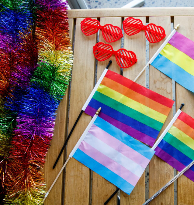 Rainbow-colored pride flags, red heart-shaped sunglasses, and rainbow tinsel on a wooden surface.