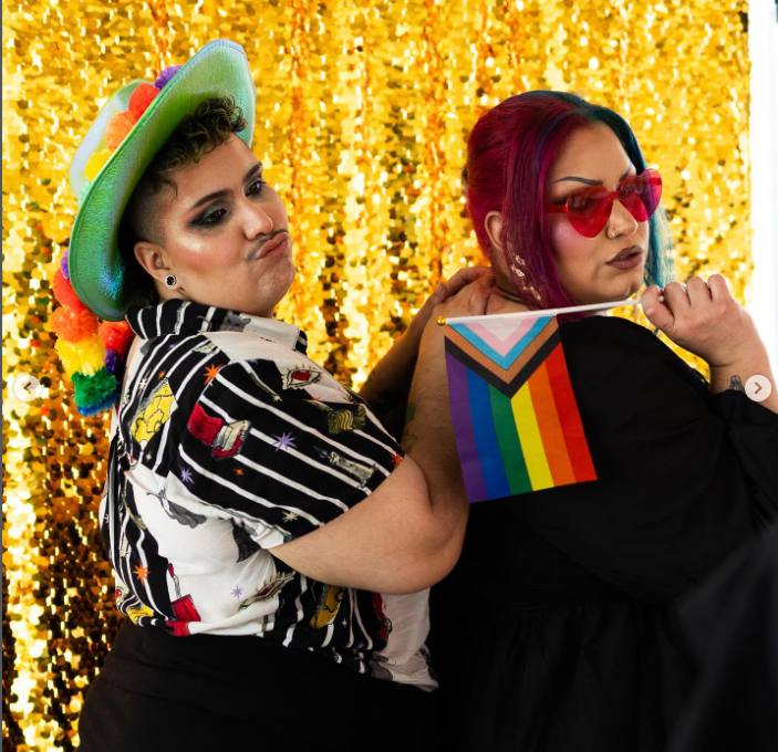 Two individuals celebrating LGBTQ Pride, one wearing a colorful hat and a patterned shirt, and the other with rainbow sunglasses and a rainbow flag against a gold sequin background.