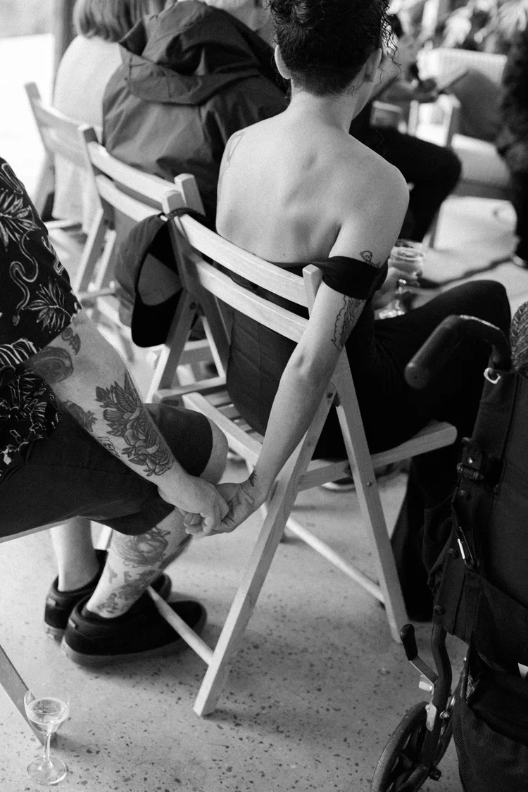 A person with tattoos on their arm is sitting on a wooden chair and holding hands with another person standing next to them. The person sitting is shirtless and has short, curly hair. The scene appears to be in a gathering or event setting, with othe